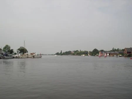 Calm water body with boats docked at shore and distant trees and buildings under overcast sky