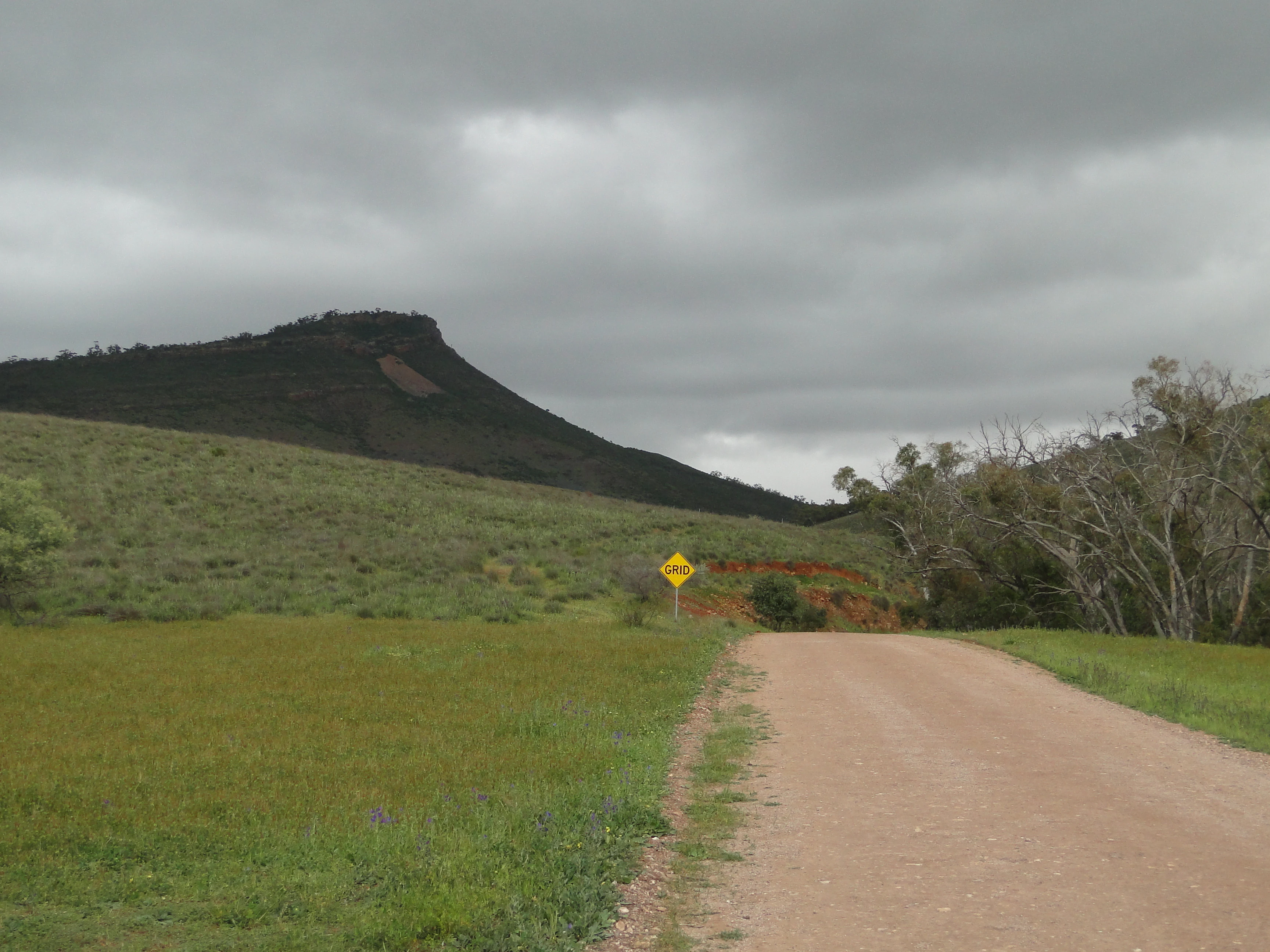 A dirt track leads through a grassy field toward a dark, rocky ridgeline under an overcast sky, with a yellow road sign visible in the midground.
