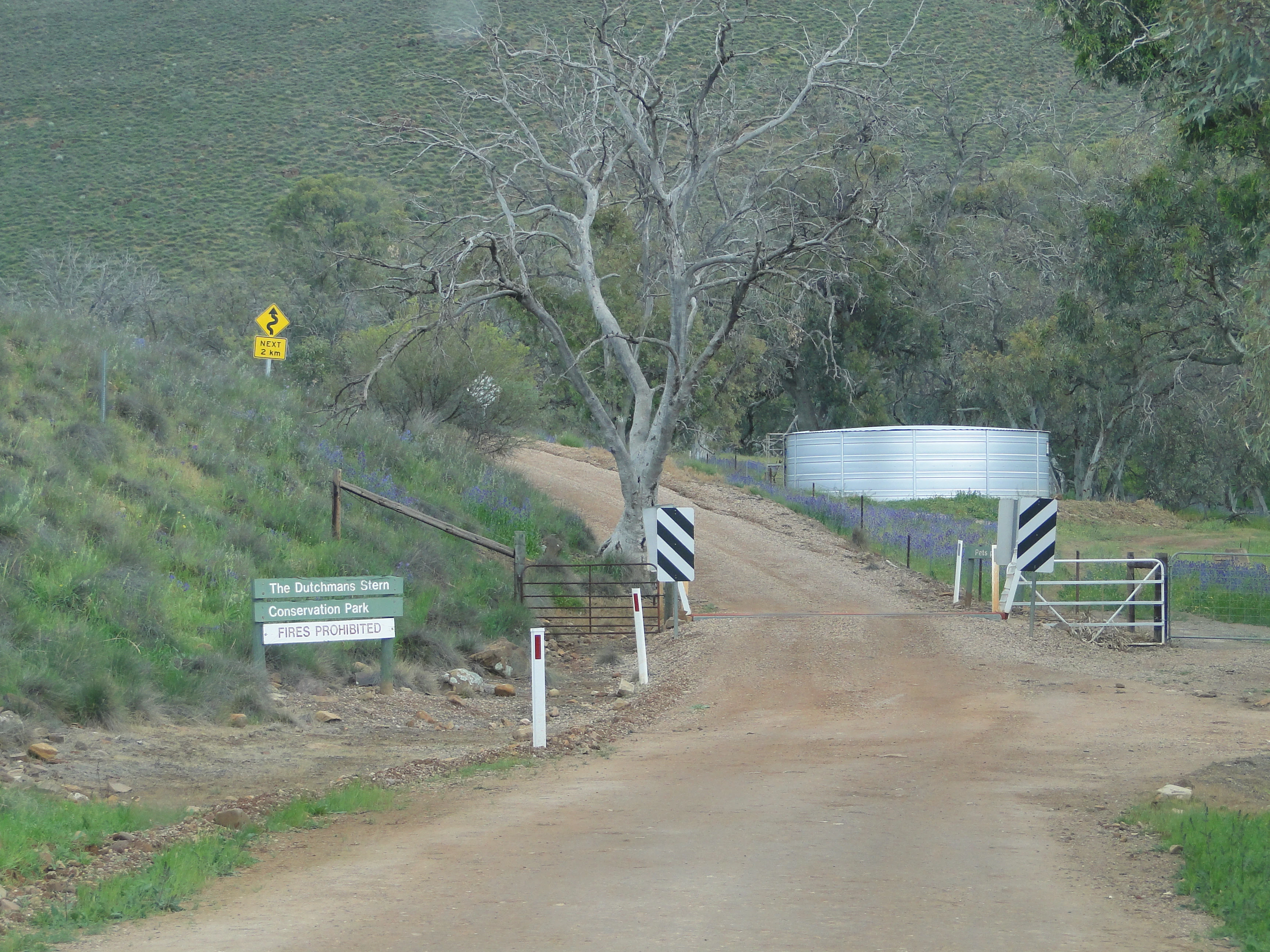 A dirt access road leading into The Dutchmans Stern Conservation Park, marked by a green sign and a metal gate structure with a corrugated water tank overhead.