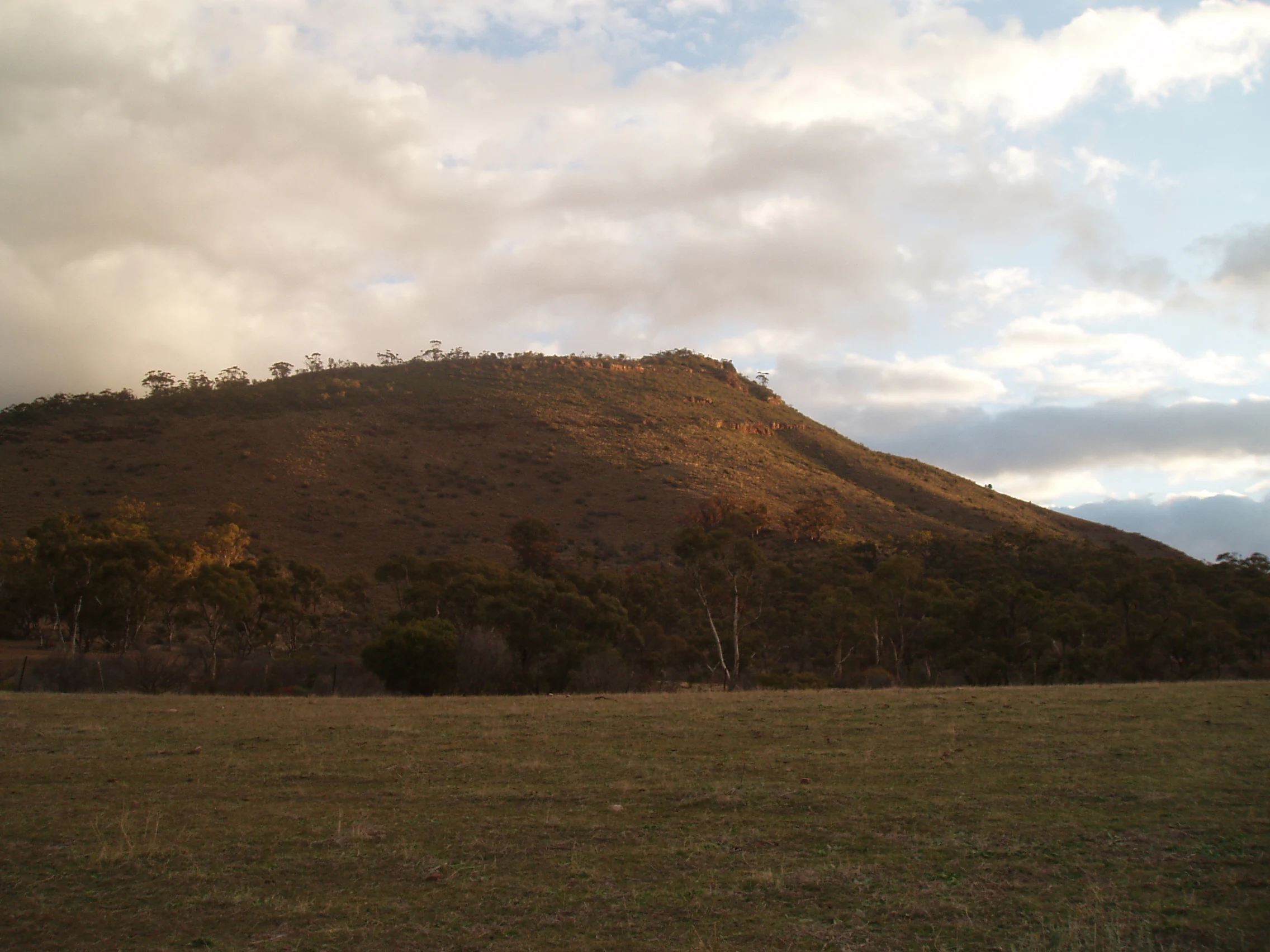 A low-angle view of a rounded, brown, sunlit hill rising above a dry, grassy field under a cloudy sky.