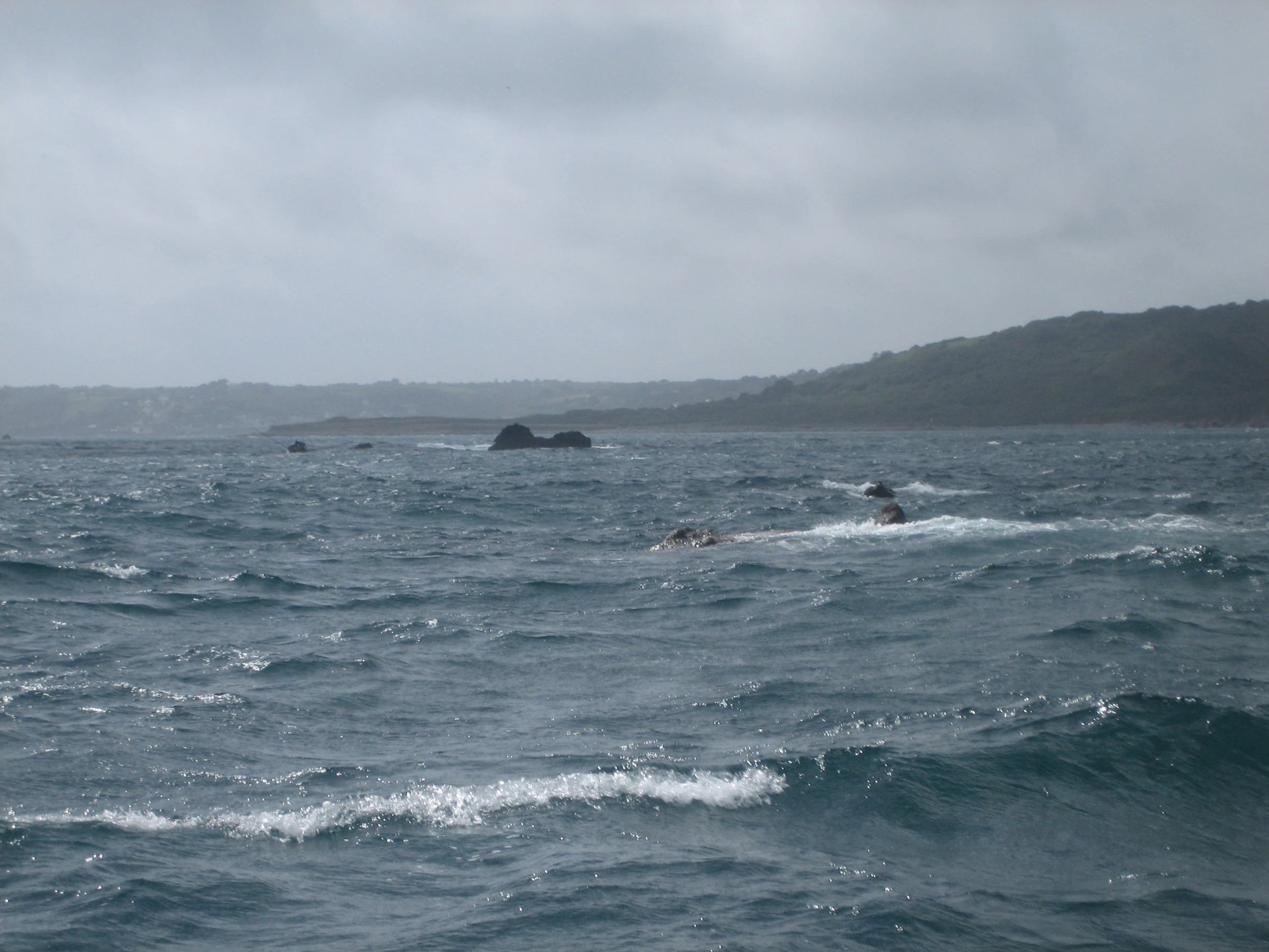 A view of the Manacles rocks and reefs protruding from the sea off a coastal headland under a cloudy sky.