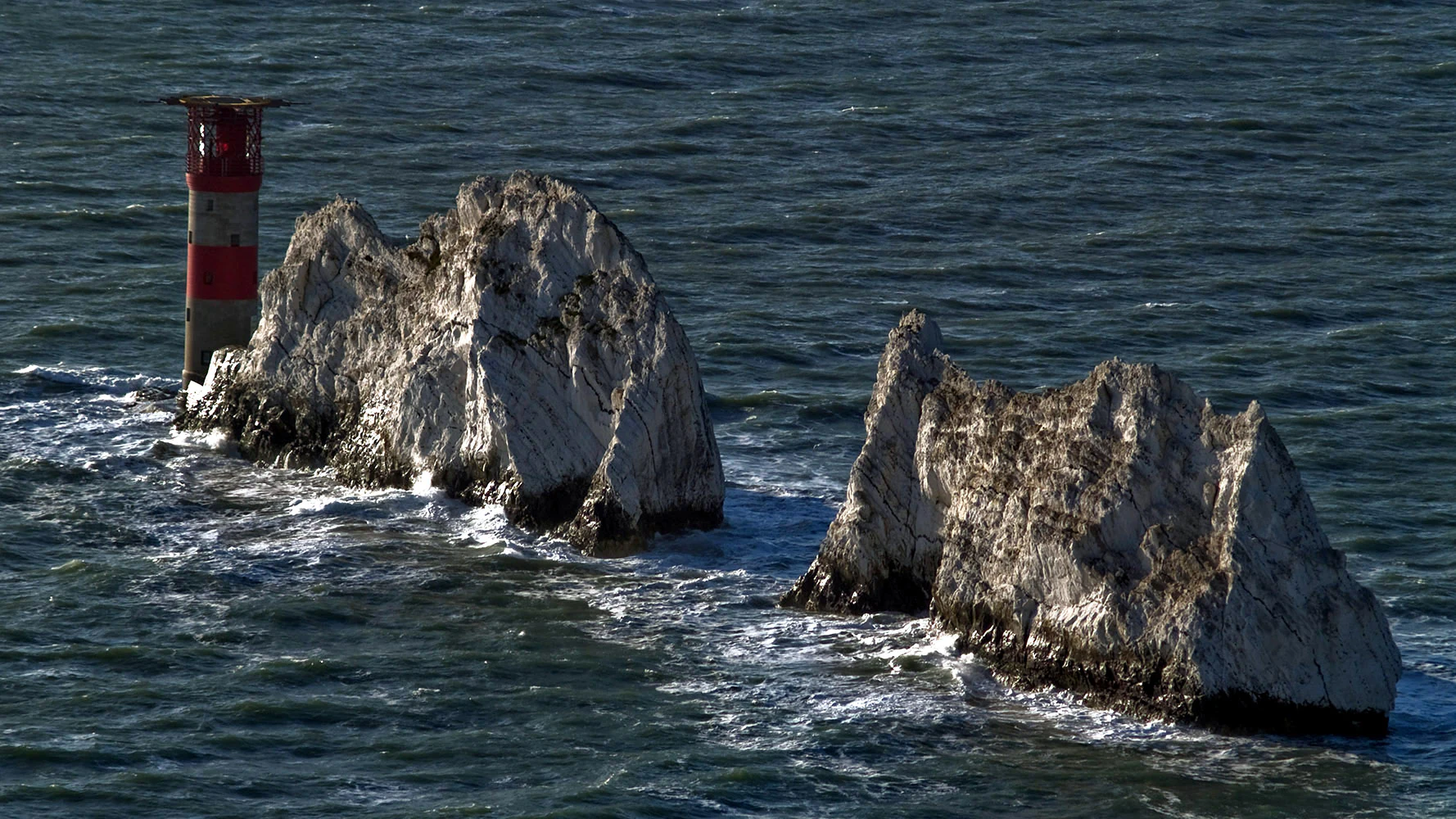 A close up aerial view of the Needles, three jagged chalk rock stacks emerging from dark sea water, with a red and white striped lighthouse standing on the edge of the nearest formation.