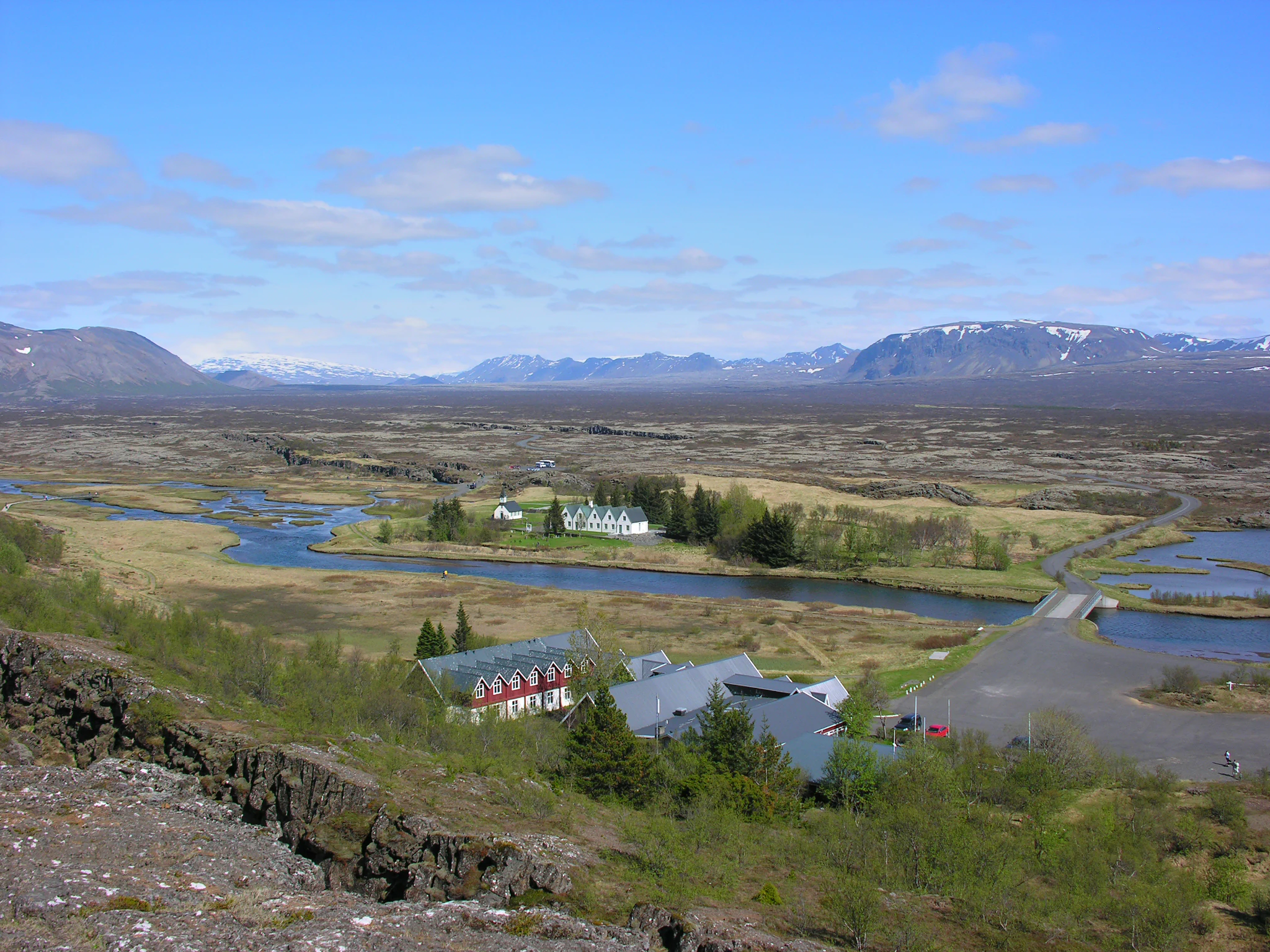 Panoramic landscape of Þingvellir National Park with rivers, grassy fields, scattered buildings, distant mountains, and clear sky