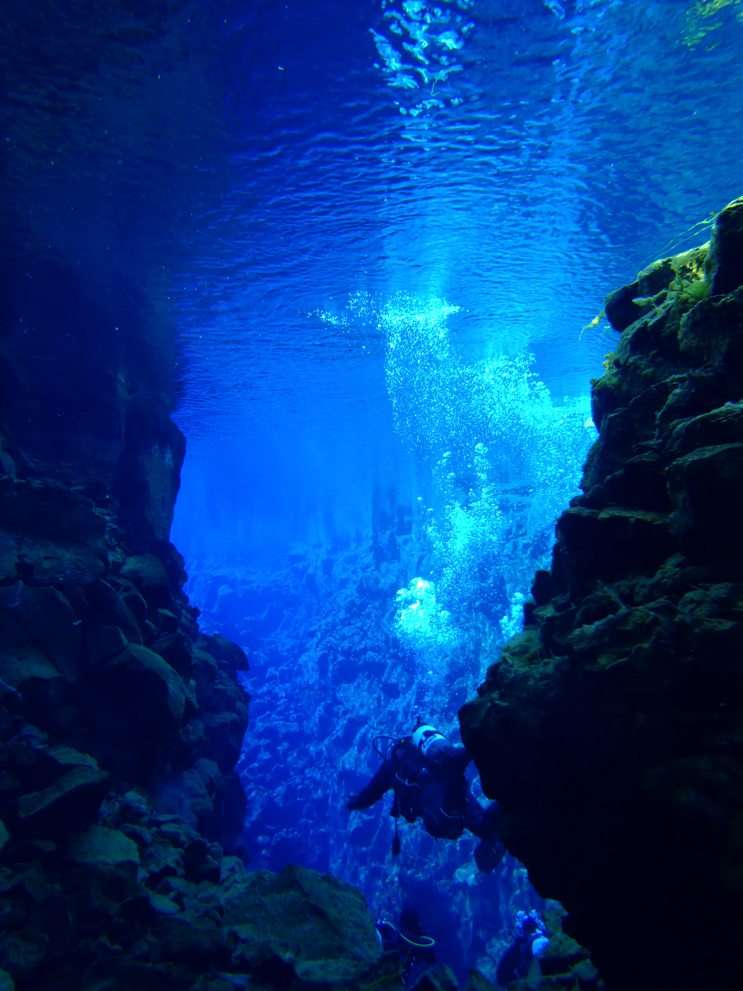 Scuba diver swimming in clear blue water between rocky walls in Silfra fissure.