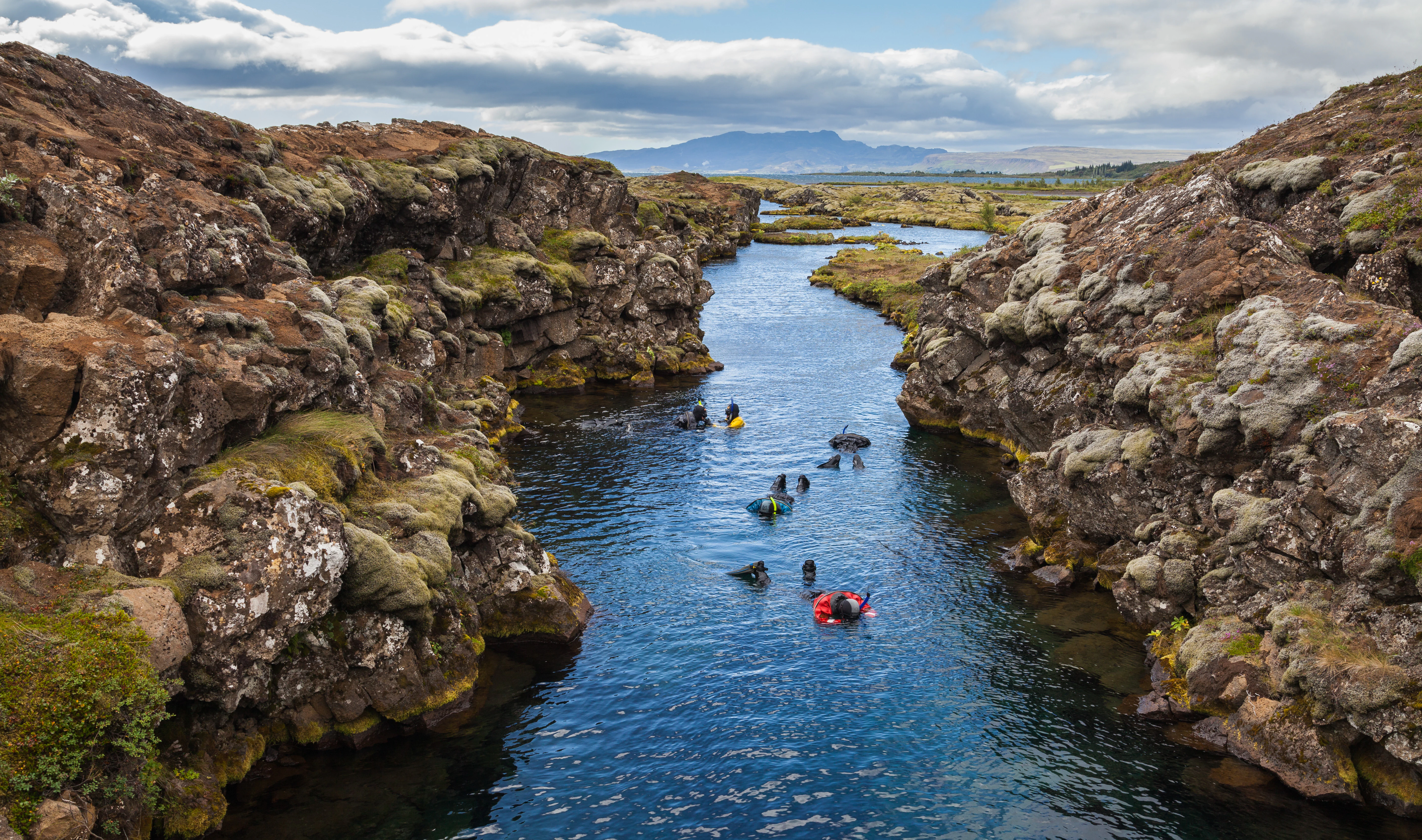 People in snorkeling gear swimming in a narrow waterway between rocky cliffs in Þingvellir National Park, Iceland