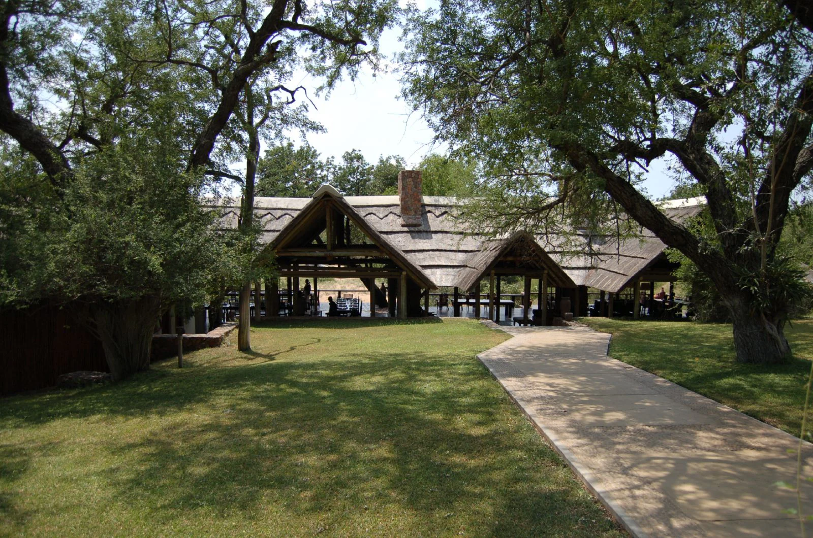 A low, open-air lodge building with a traditional thatched roof, surrounded by mature trees and a manicured lawn, approached by a paved walkway.