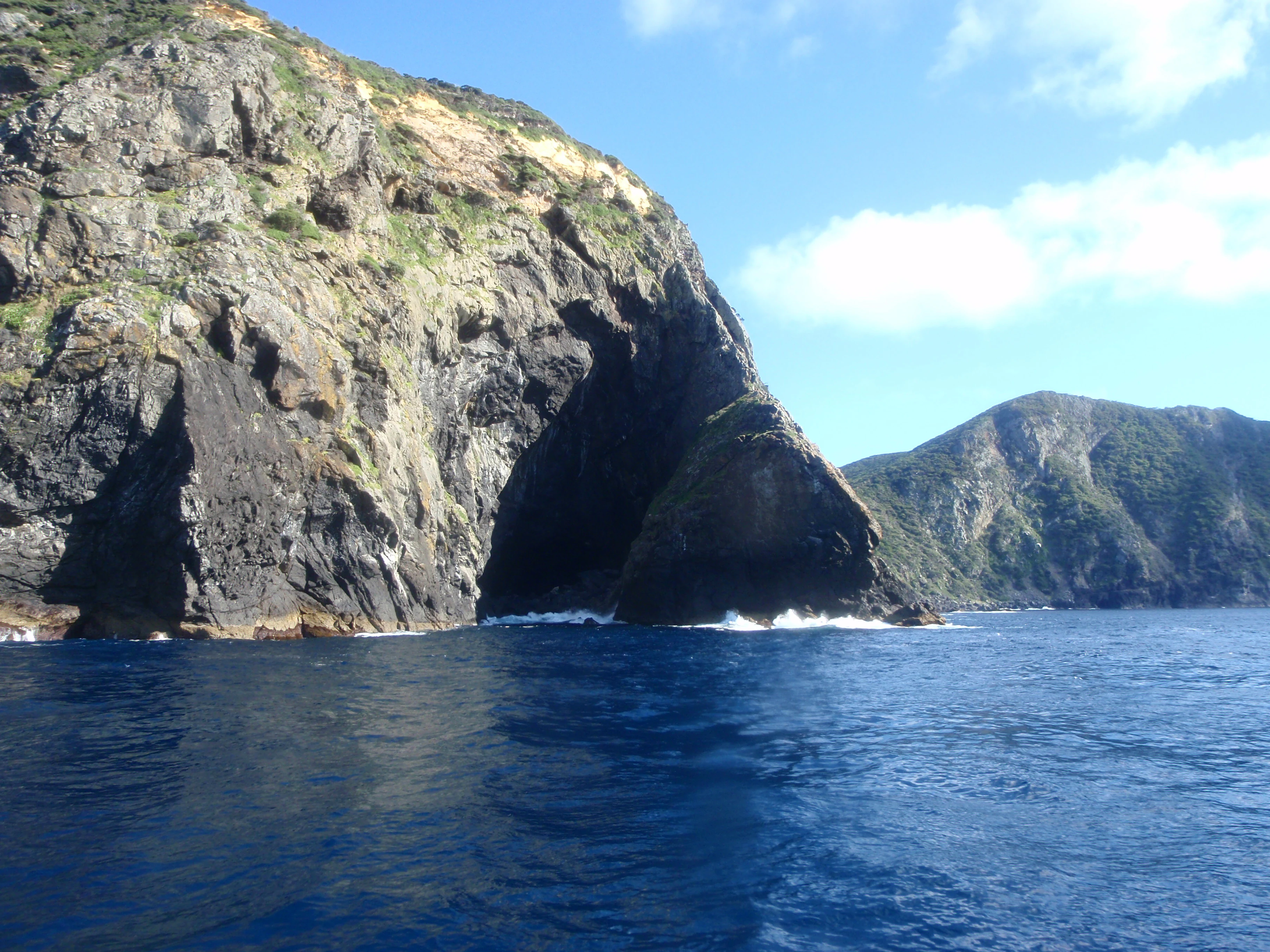 A view from the water showing dramatic, steep rocky cliffs of the Three Kings Islands rising directly from the ocean, with a natural cave formation visible in the base of the primary cliff.