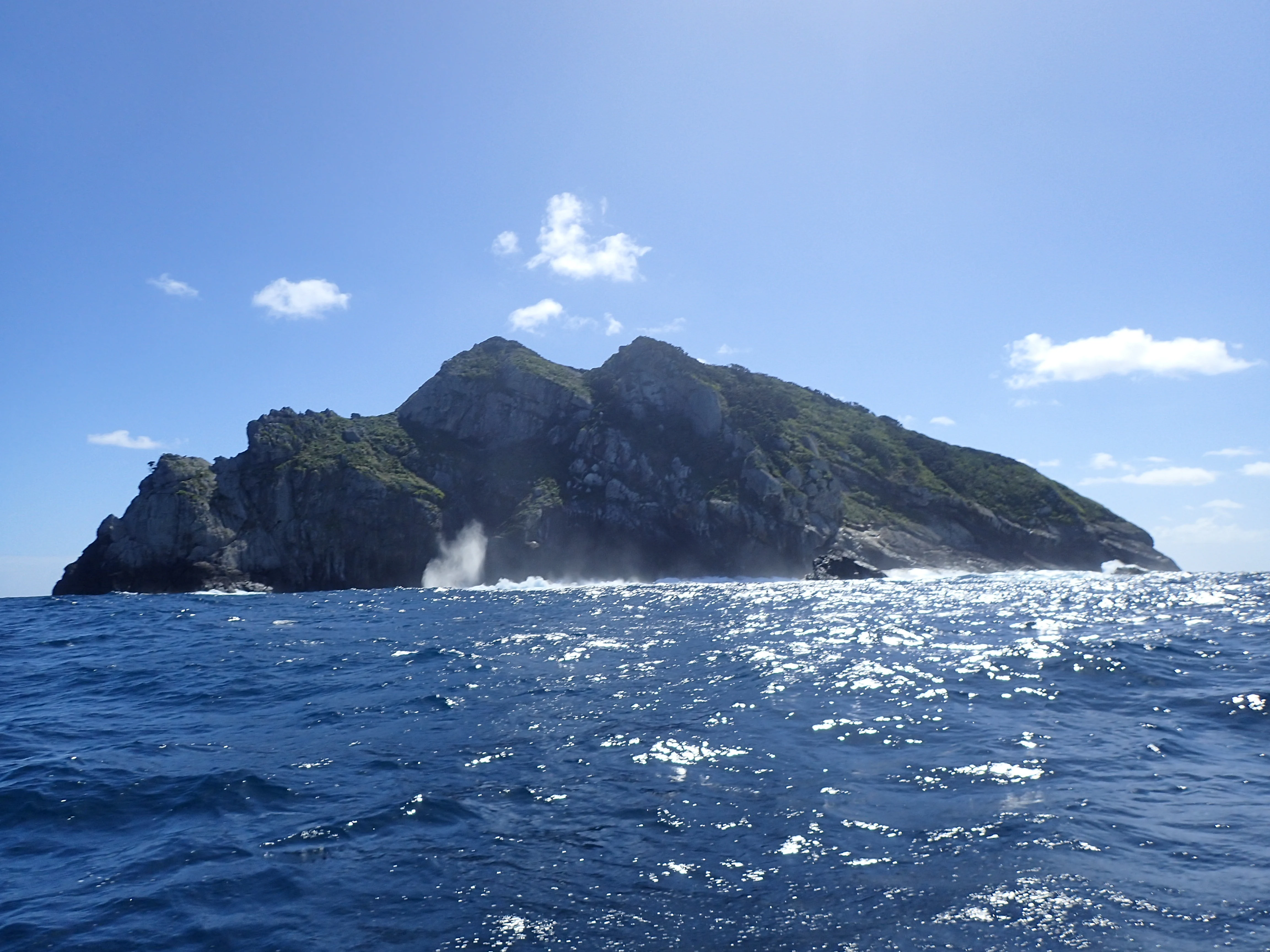 A rugged island rising from the deep blue ocean with steep, rocky cliffs and visible surf breaking against its base under a clear sky.