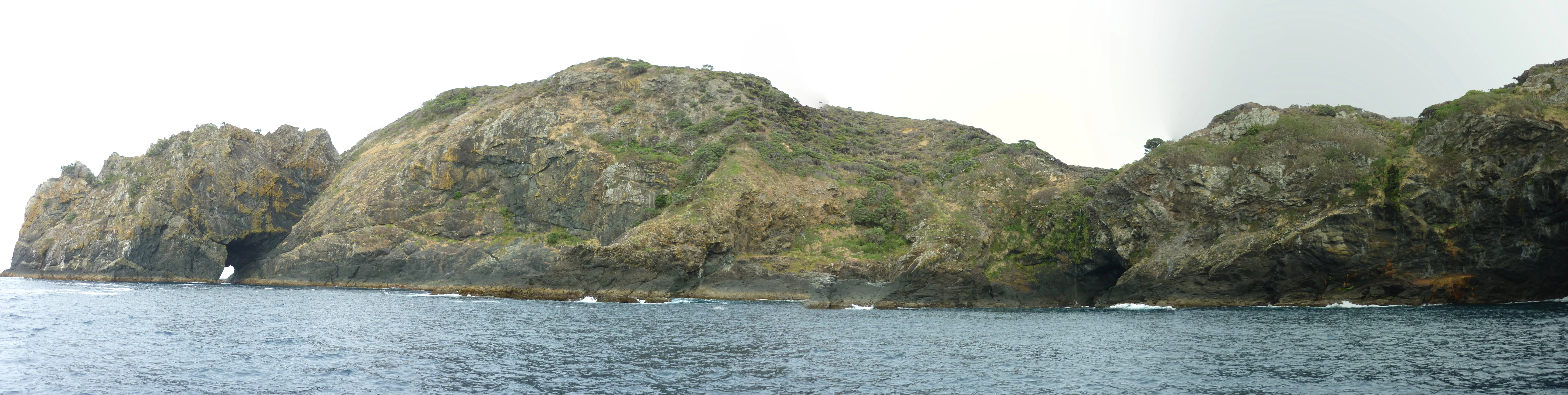 A panoramic view of a rocky, green, and hilly island coastline rising directly out of the deep blue ocean, featuring small sea caves and cliffs.