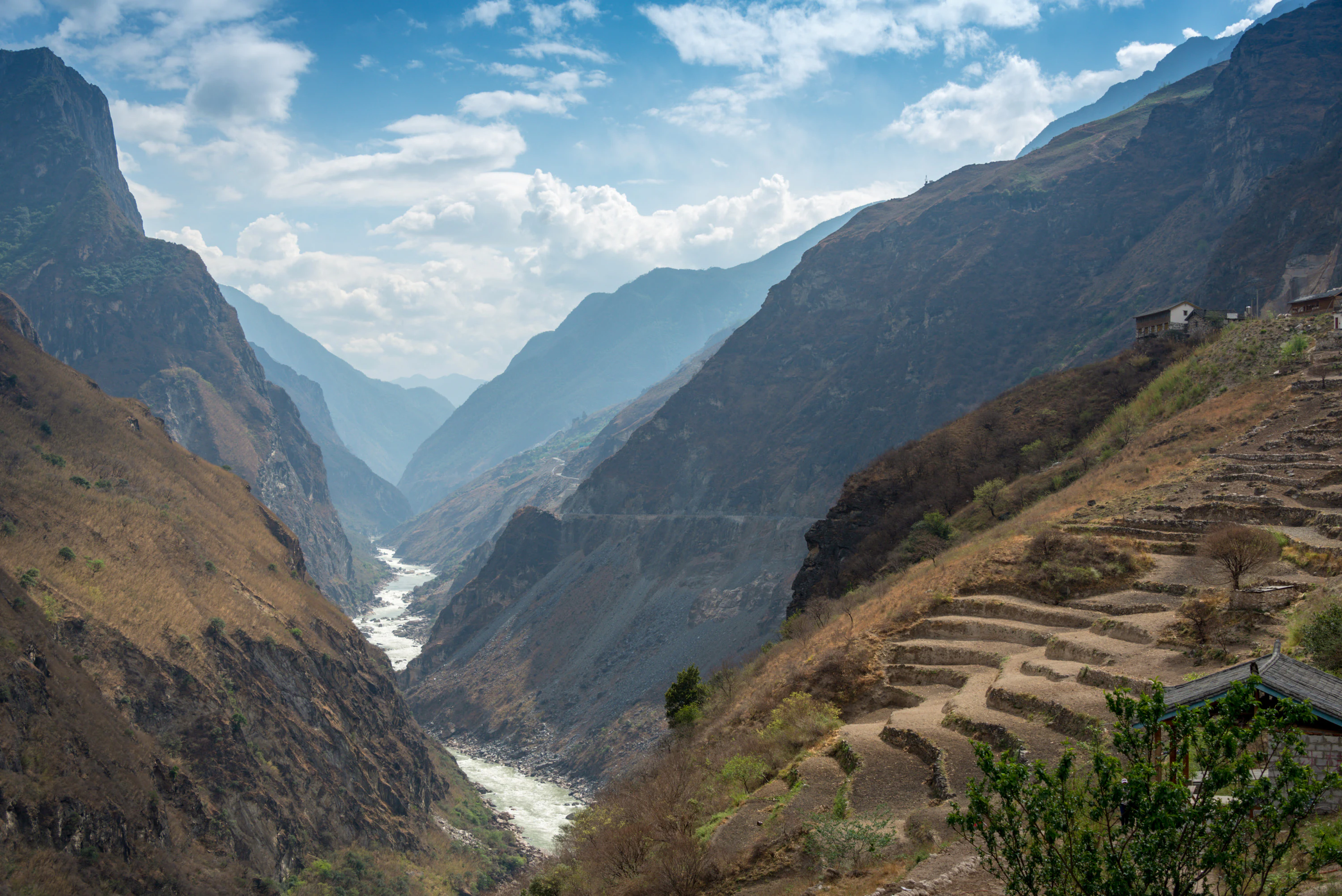 A wide canyon landscape with a winding river flowing between steep, rugged mountain slopes, featuring terraced agricultural land on the right side.