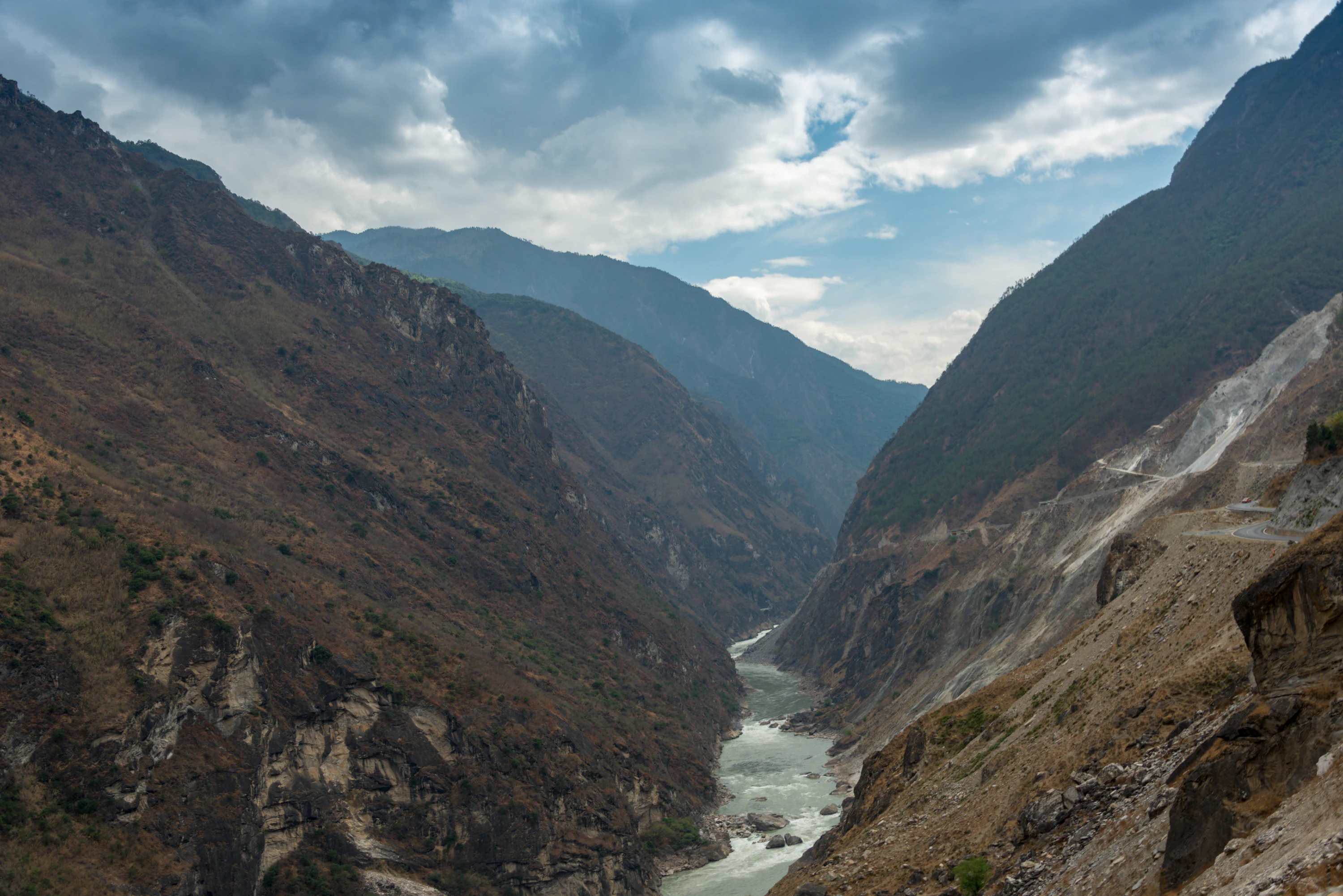 A wide angle shot looking down a steep, rocky canyon with a river flowing through the center. Large, rugged mountains rise on both sides under a cloudy sky.