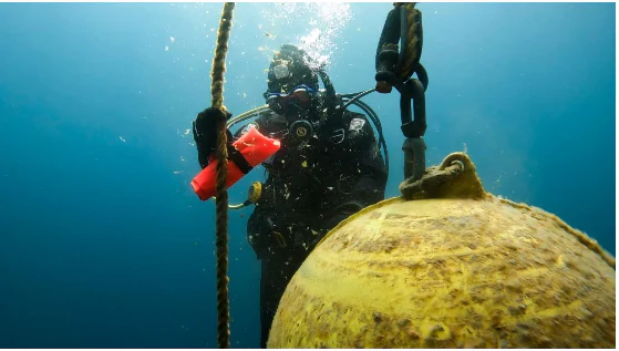 Una vista submarina de un buceador con equipo completo sujetando una cuerda, junto a una gran boya de amarre amarilla, con agua azul y clara a su alrededor.