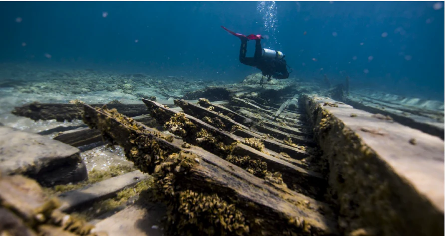 Vista submarina de un buceador explorando las nervaduras estructurales de madera de un naufragio en el fondo del lago.