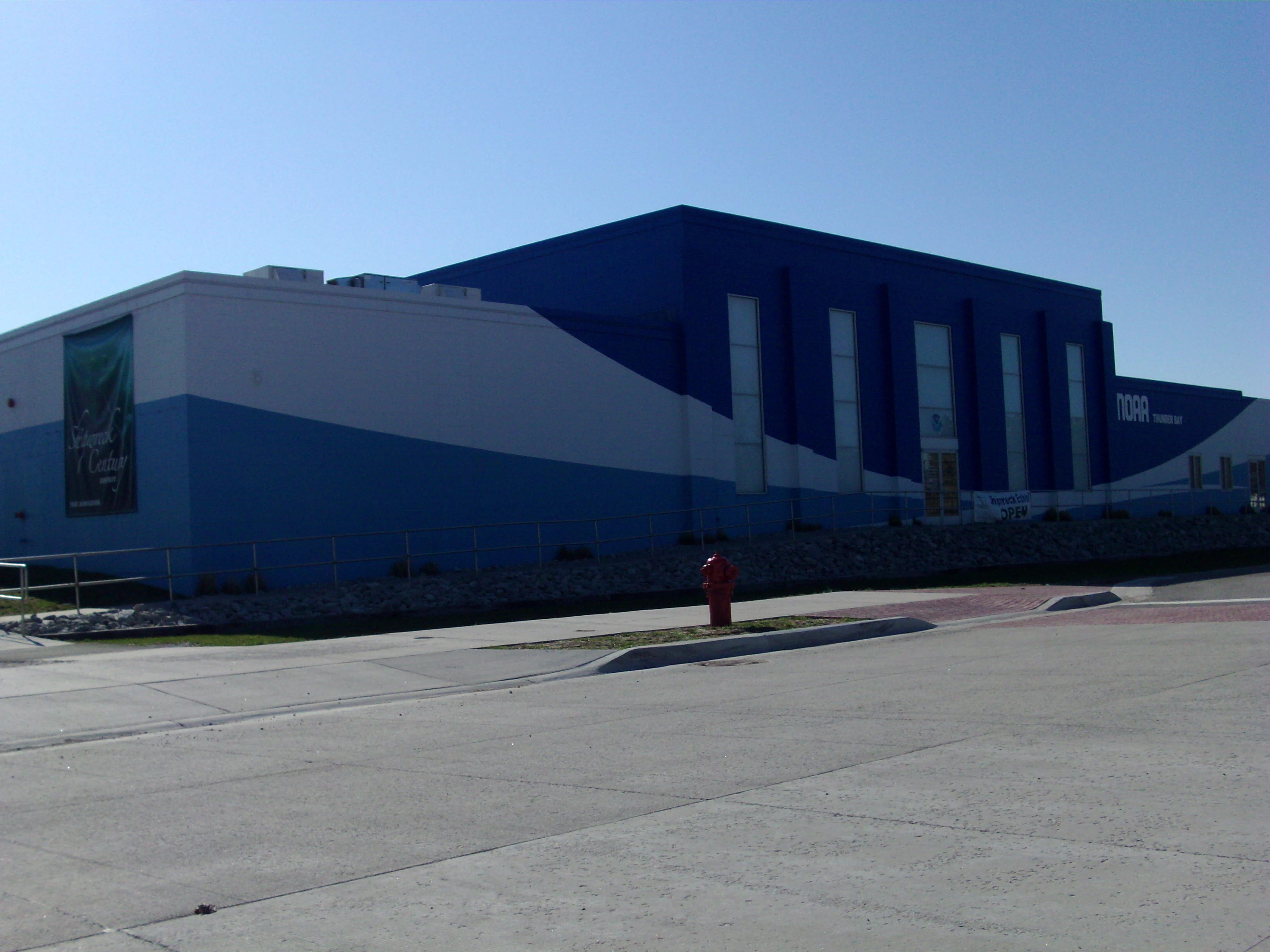 Una vista exterior del edificio del Centro Marítimo de los Grandes Lagos en Alpena, Míchigan, con paredes exteriores azules y blancas y un cielo azul despejado.