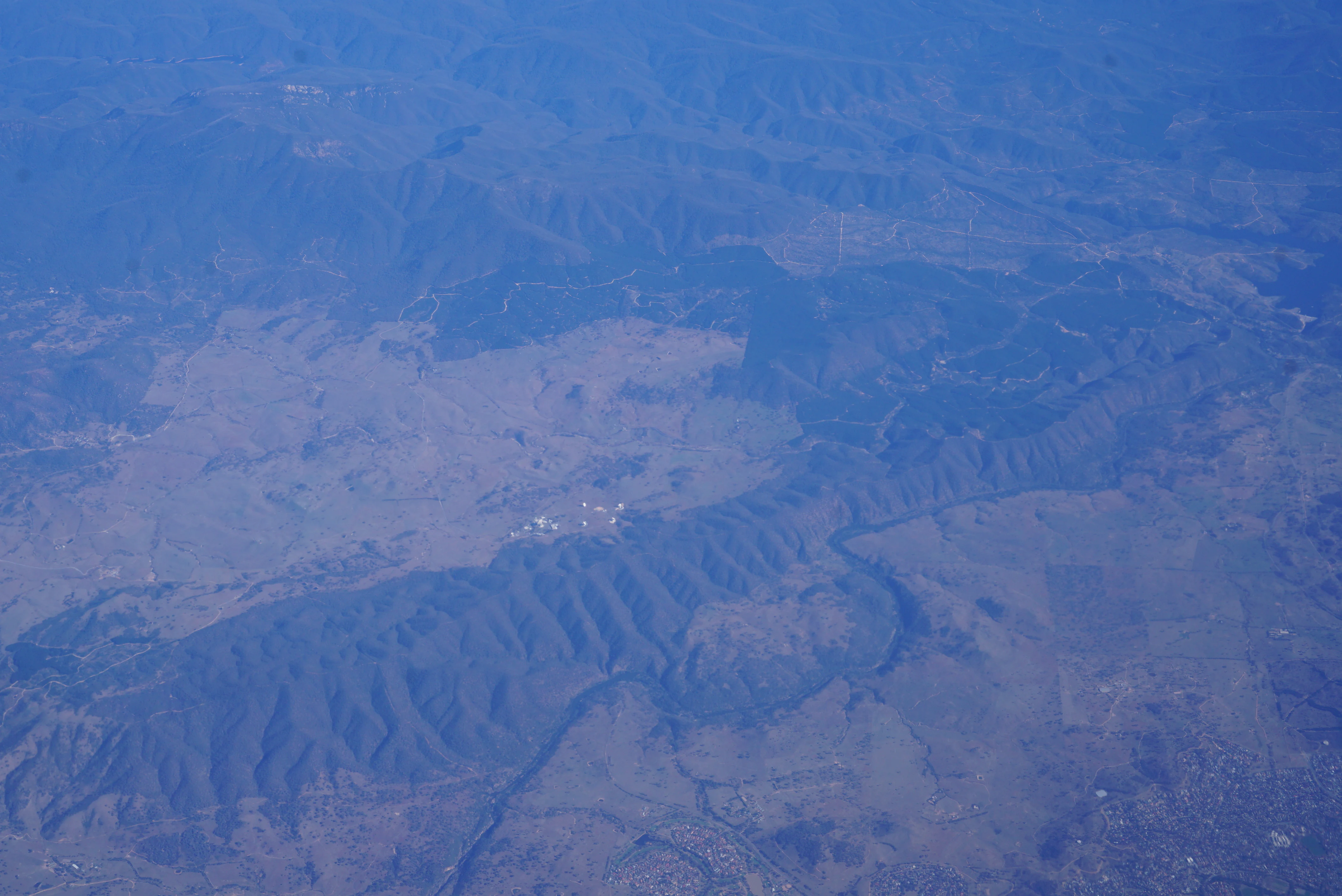 High altitude aerial view of the Australian landscape featuring mountain ridges, forested areas, and the white antennae of the Canberra Deep Space Communication Complex.