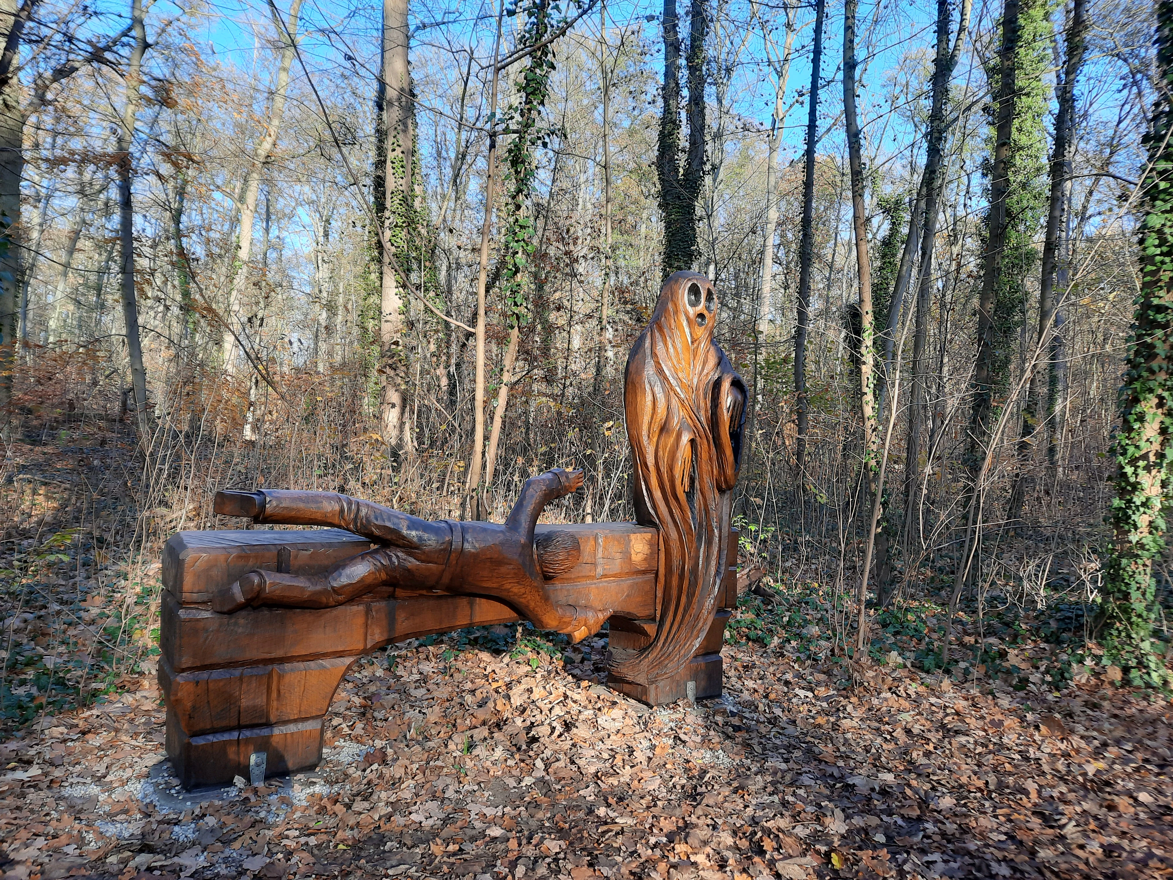 A brown wooden sculpture resembling a draped ghost or figure carved into a block, set in a wooded area with many bare trees and dry leaves on the ground.