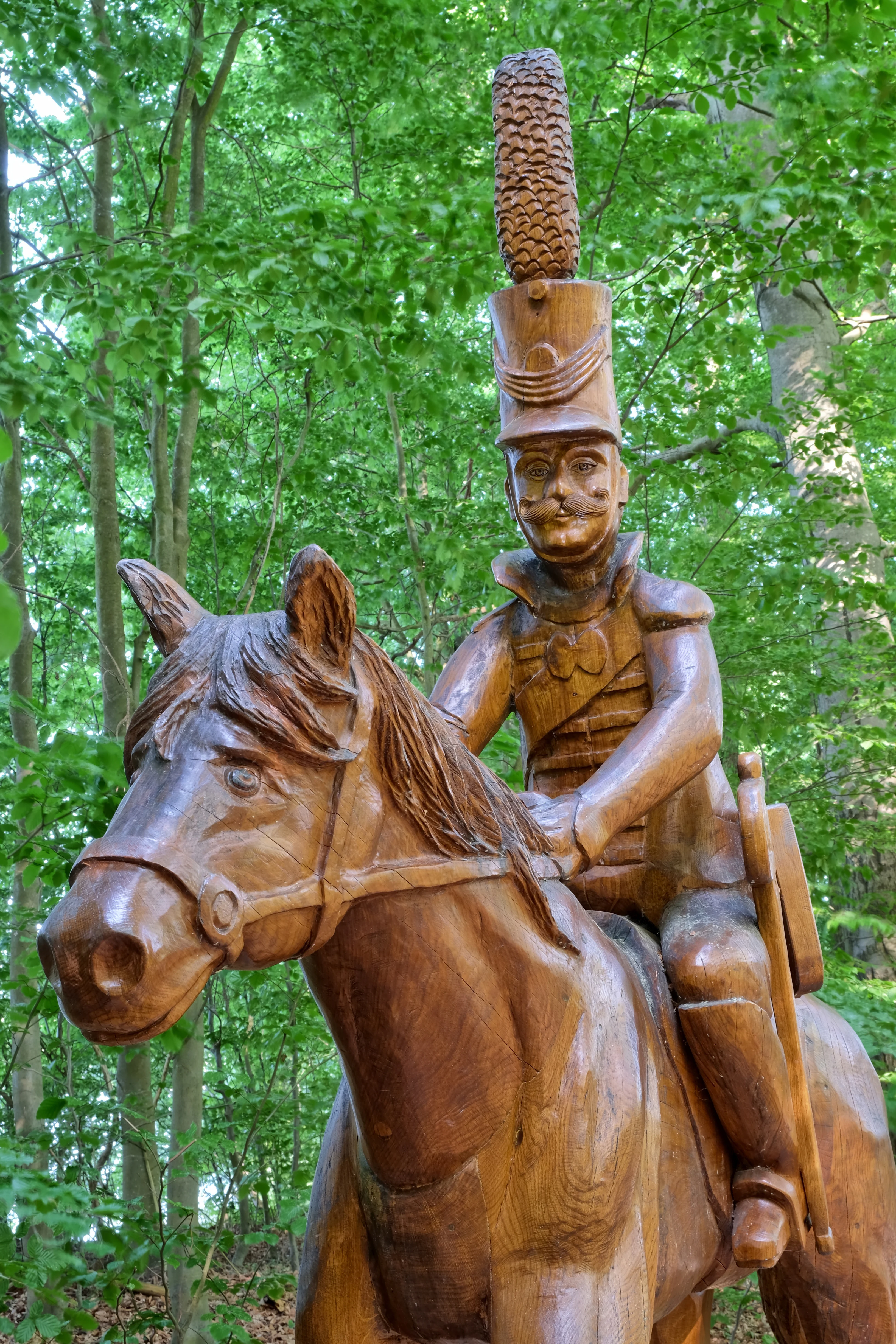 A close up view of a carved wooden statue of a hussar in a tall shako hat riding a horse, set against a background of lush green deciduous forest trees.
