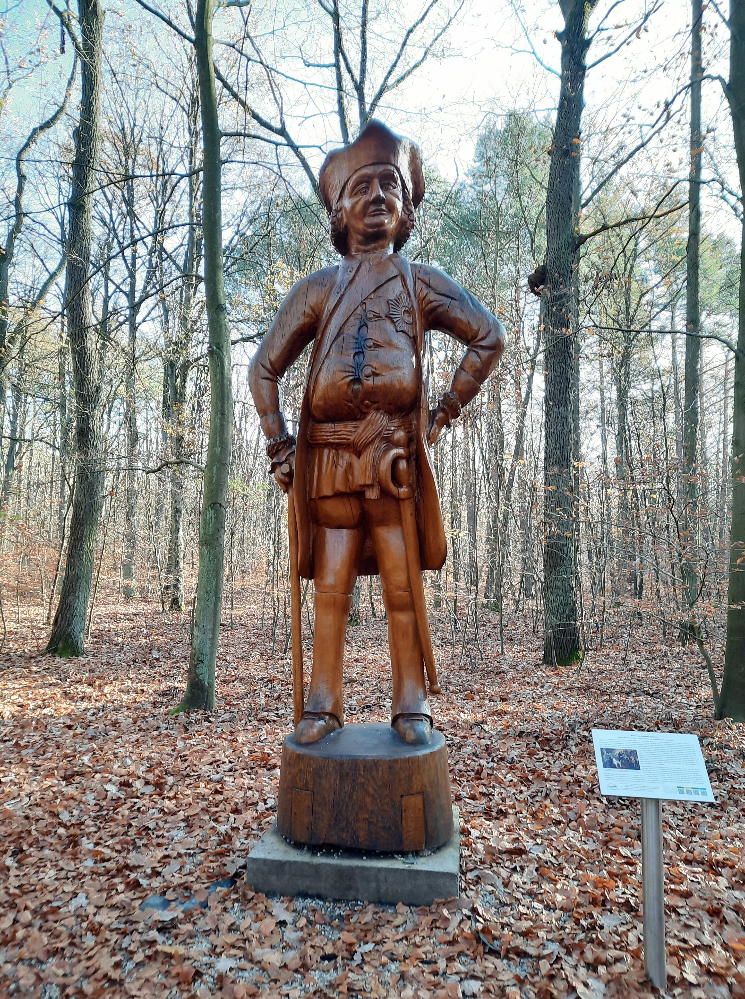 A carved wooden statue of a man in 18th-century military attire, standing on a round wooden base in a forest. To the right, an information sign on a metal post is visible.