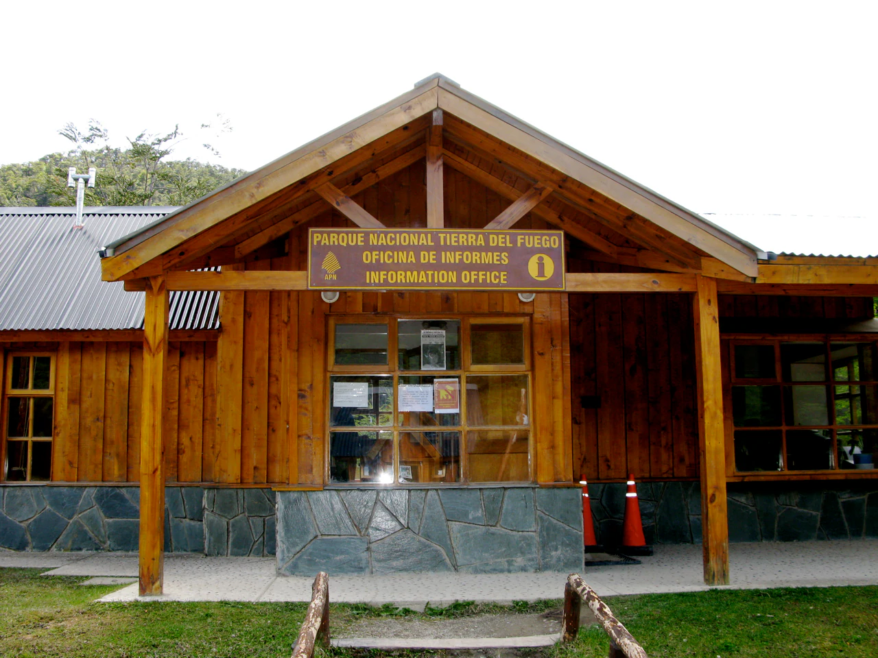 Wooden building with stone base, sign reading 'Parque Nacional Tierra del Fuego Oficina de Informes' above entrance, two orange traffic cones on concrete path