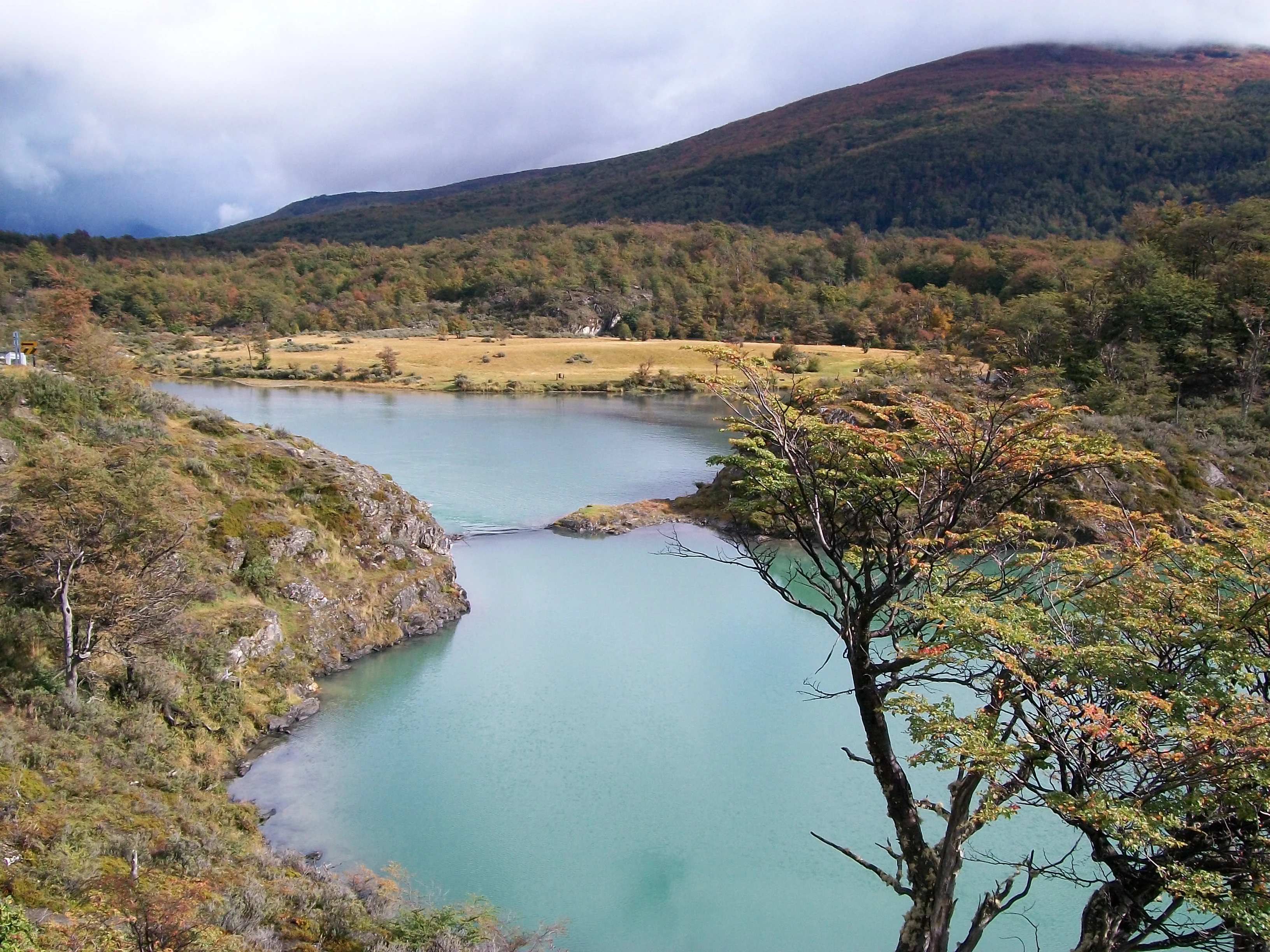 A turquoise river winds through a landscape of rocky hills and autumn-colored trees, with forested mountains in the background under a cloudy sky