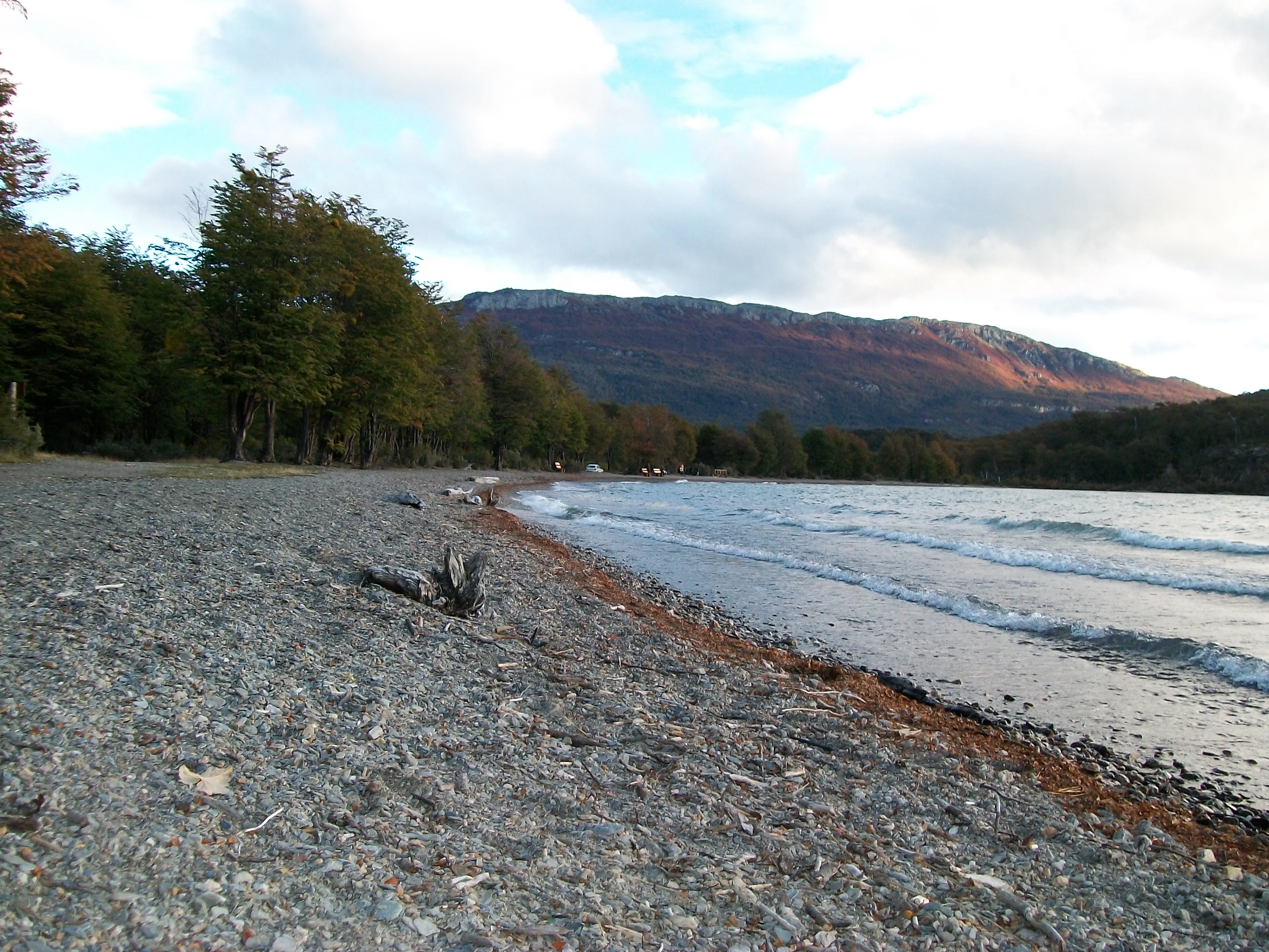 Gravel beach along Lago Roca with forested hills and distant mountains under a partly cloudy sky