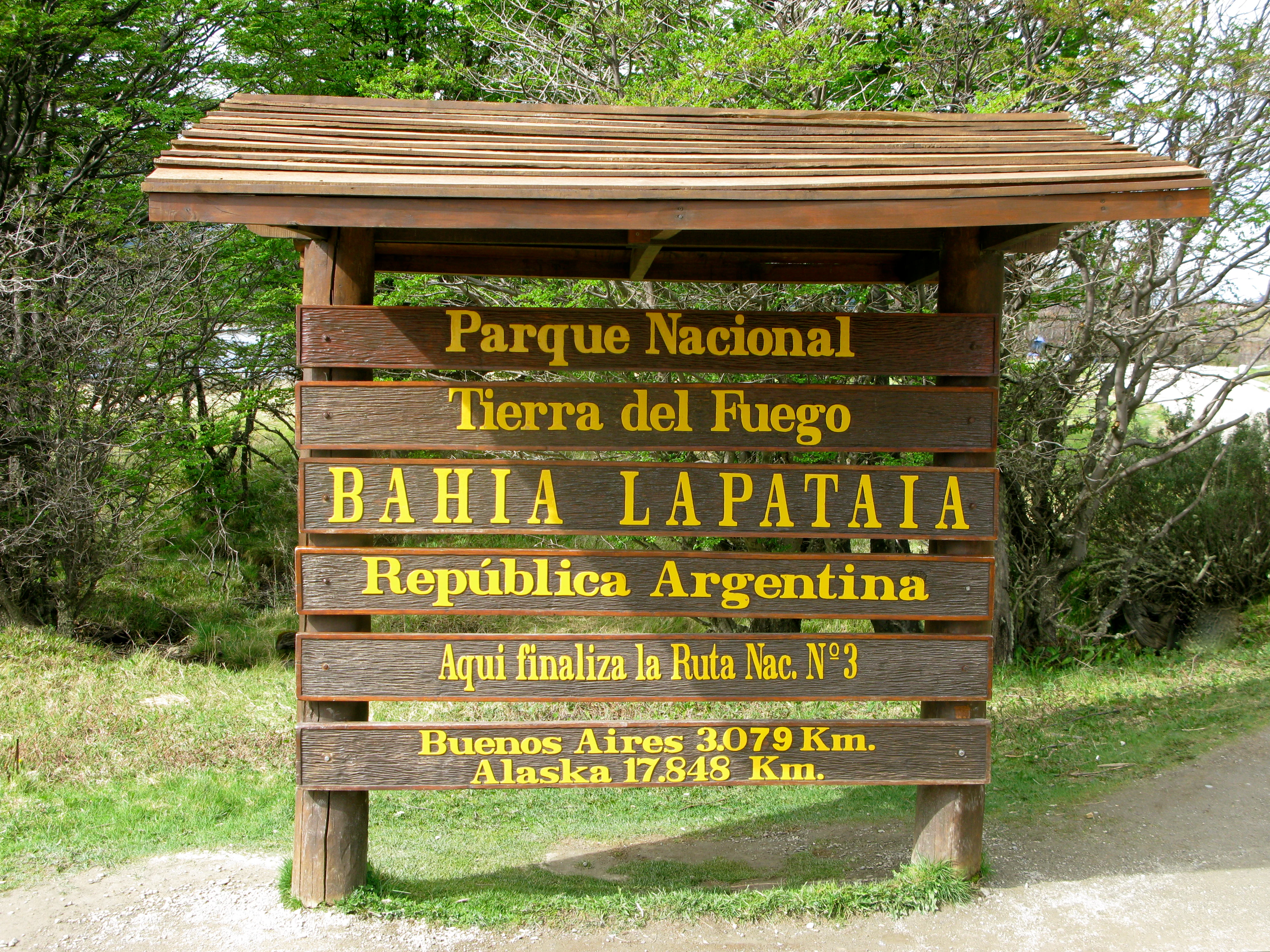 Wooden sign structure with multiple plaques displaying 'Parque Nacional Tierra del Fuego', 'Bahia Lapataia', 'República Argentina', 'Aqui finaliza la Ruta Nac. N°3', 'Buenos Aires 3079 Km', 'Alaska 17348 Km' in yellow text on brown planks, surrounded by green grass and trees