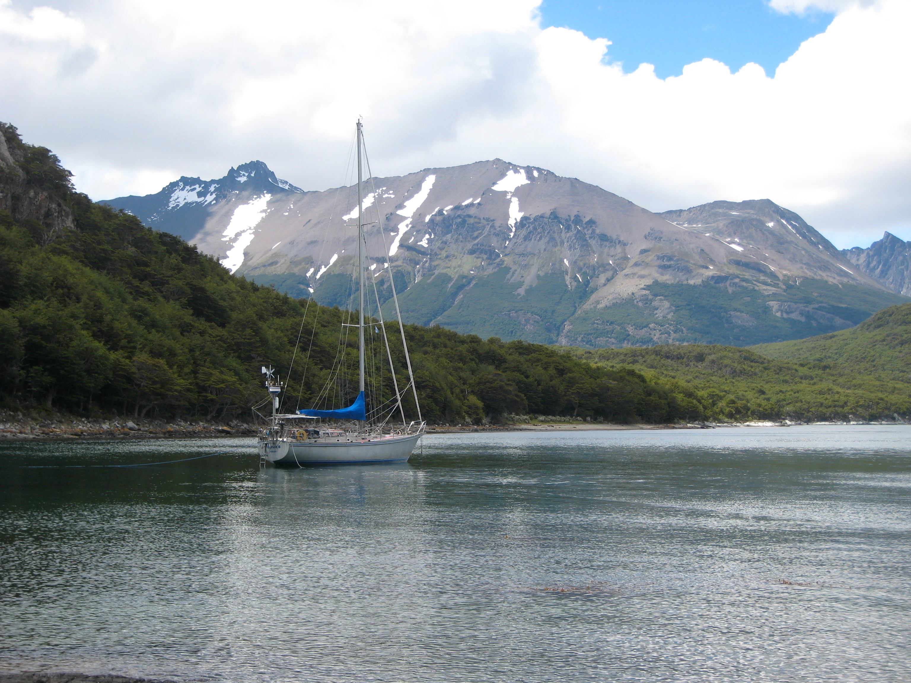 Sailboat floating on calm water with forested hills and snow-capped mountains in the background under a partly cloudy sky