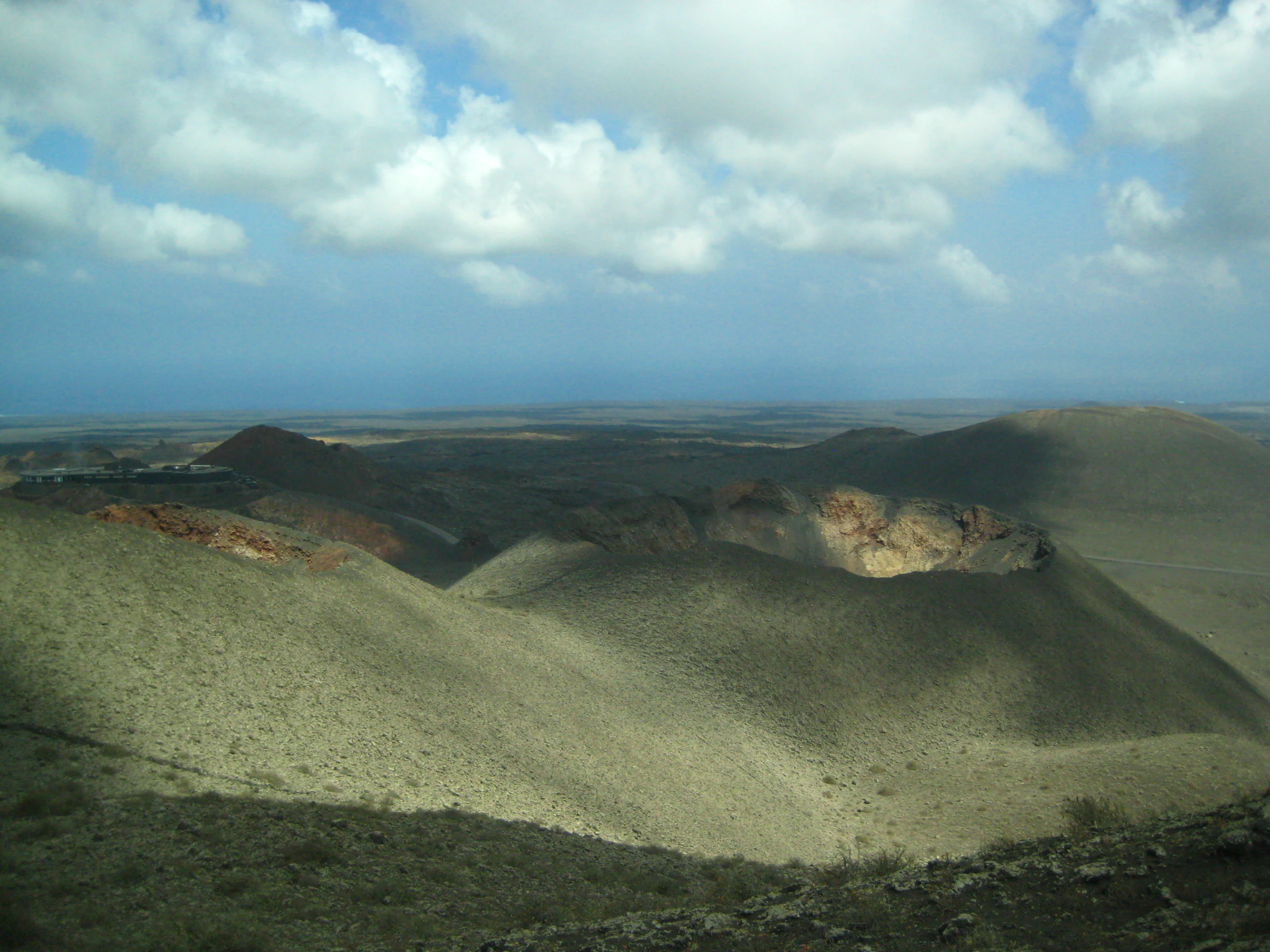 A wide landscape view of Timanfaya National Park showing volcanic hills, a crater, and open terrain under a partly cloudy sky