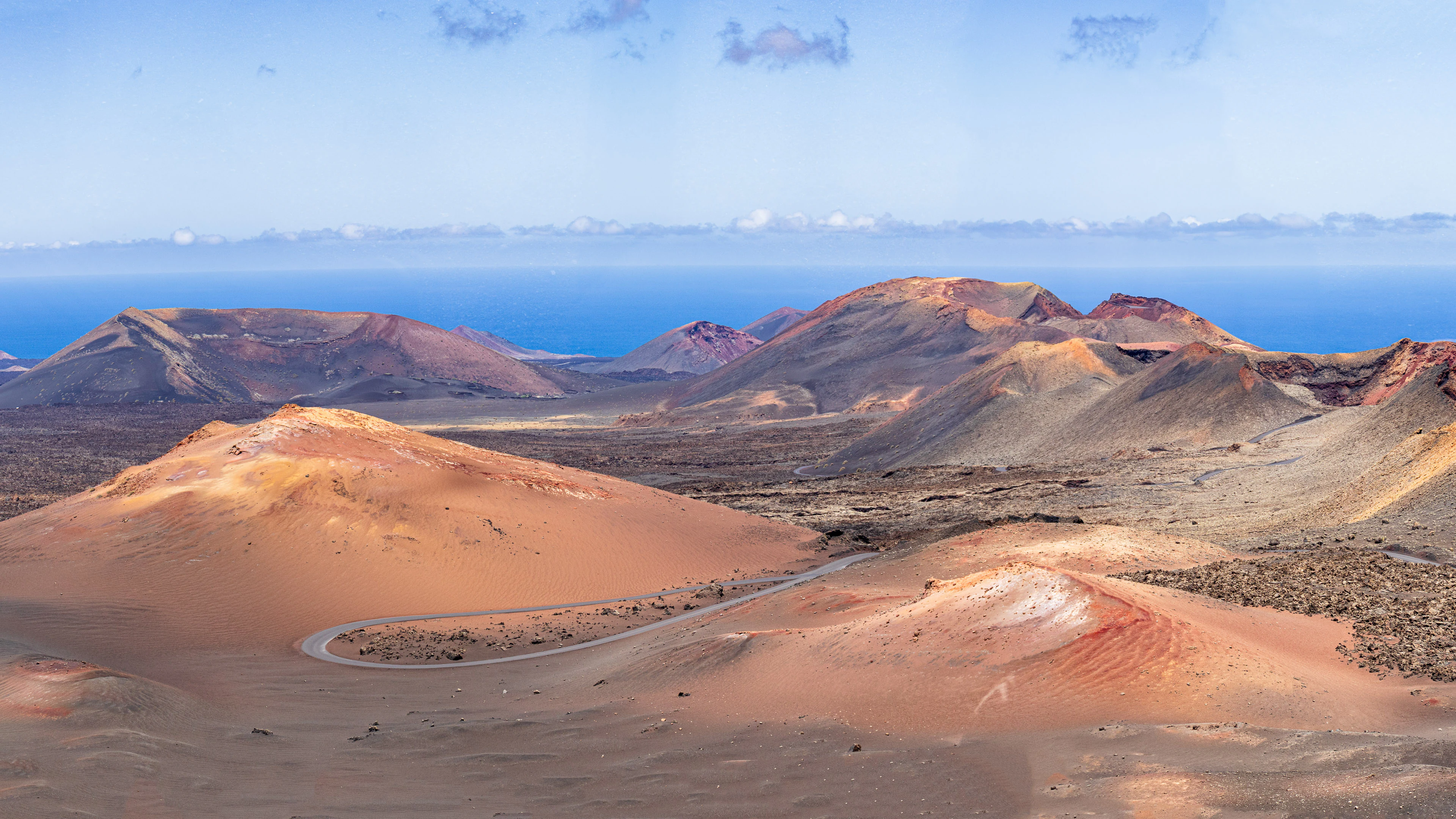 Wide landscape view of Timanfaya National Park showing red and brown volcanic hills, a winding road, and the ocean under a clear blue sky