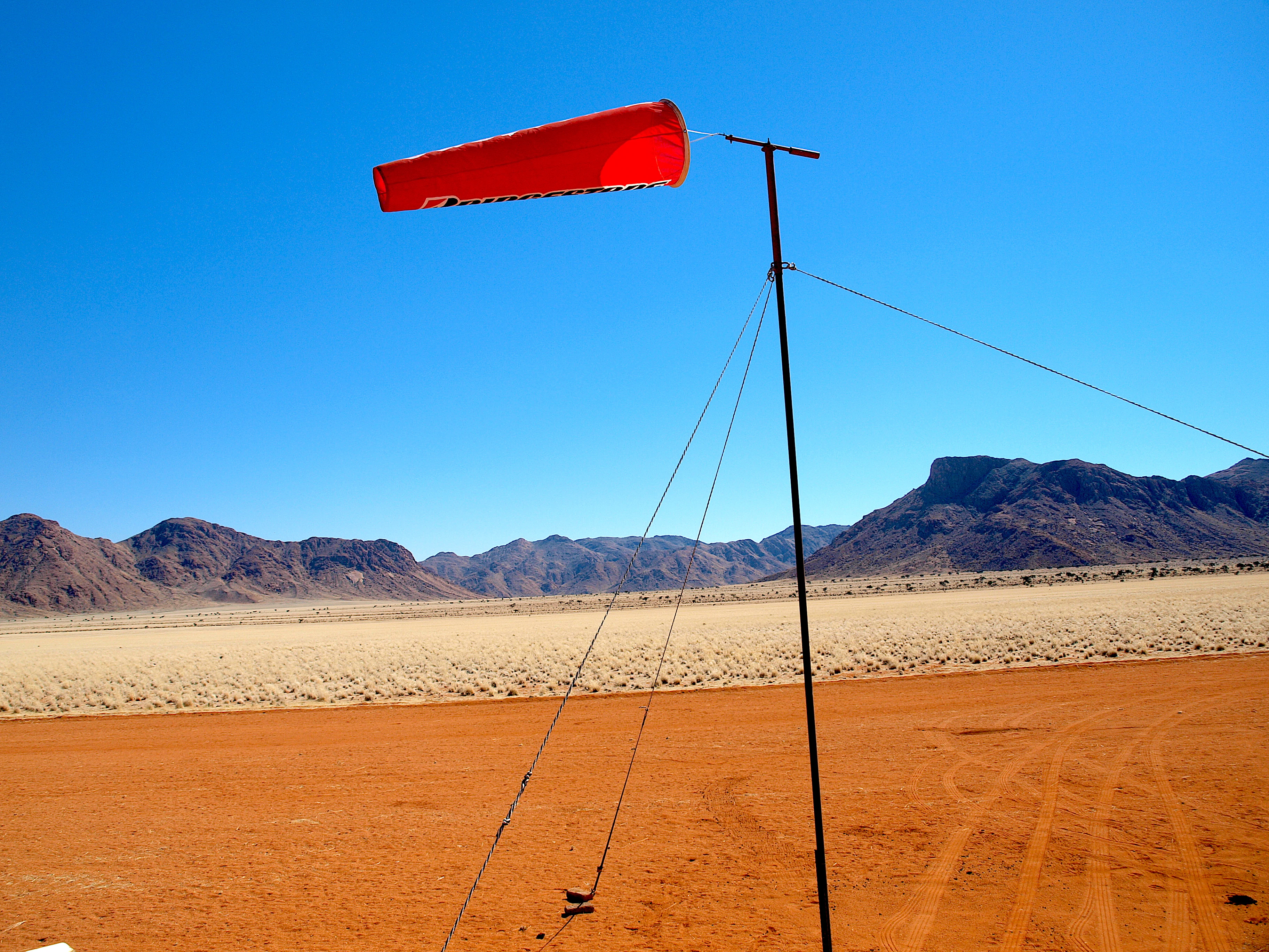 An orange windsock on a thin metal pole stands in the foreground, with an arid desert landscape and distant rocky mountains behind it.