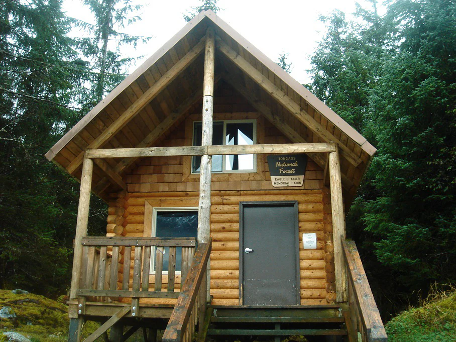 A rustic wooden cabin with a gabled roof set among dense evergreen trees, featuring a sign identifying it as a National Forest facility.