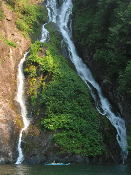 A lone kayaker floats in dark water at the base of a tall, split waterfall cascading down a steep, mossy rock cliff.