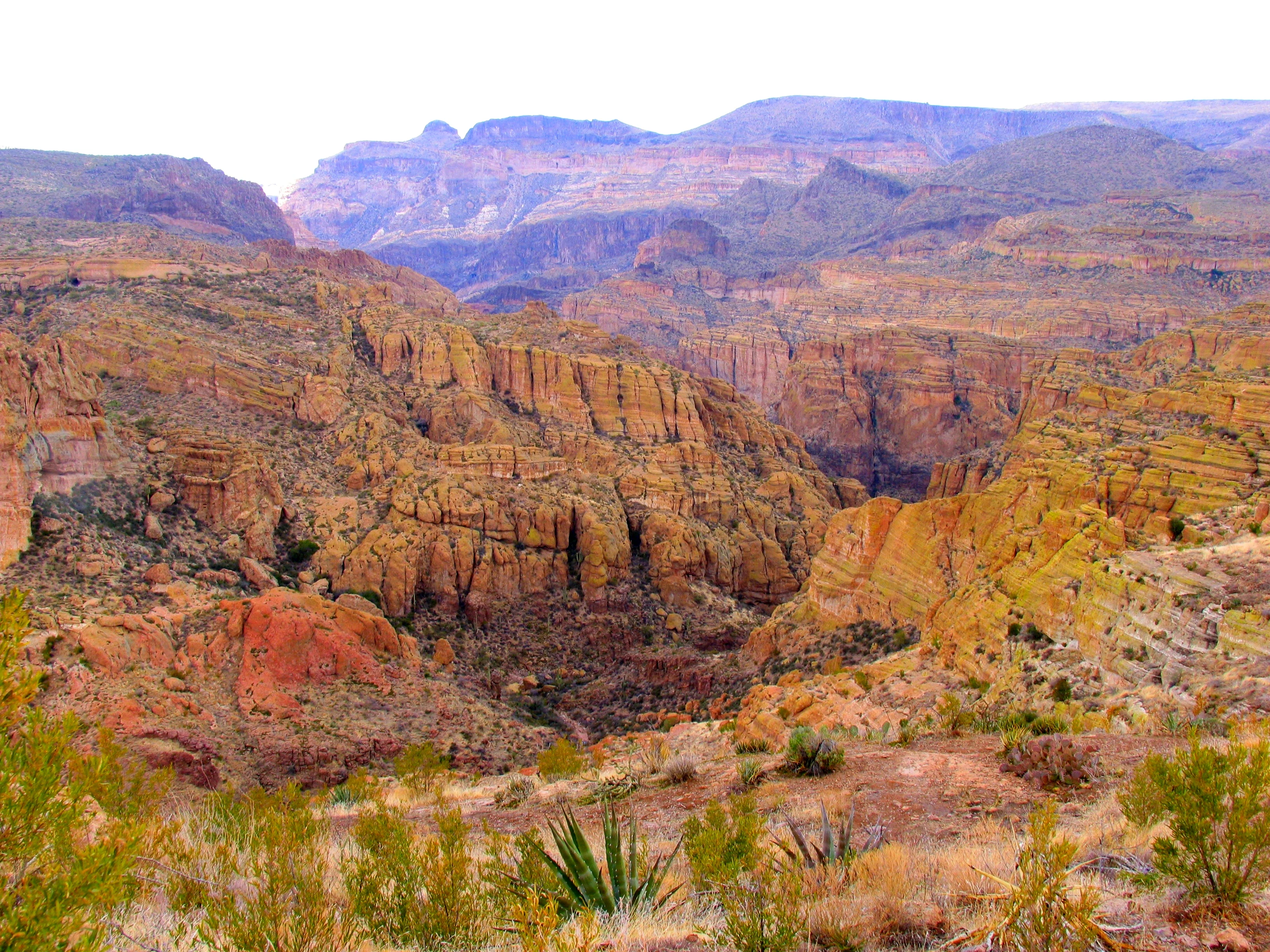Un amplio paisaje de cañón árido con formaciones rocosas estratificadas, vegetación desértica escasa y cadenas montañosas lejanas bajo un cielo brillante y nublado.