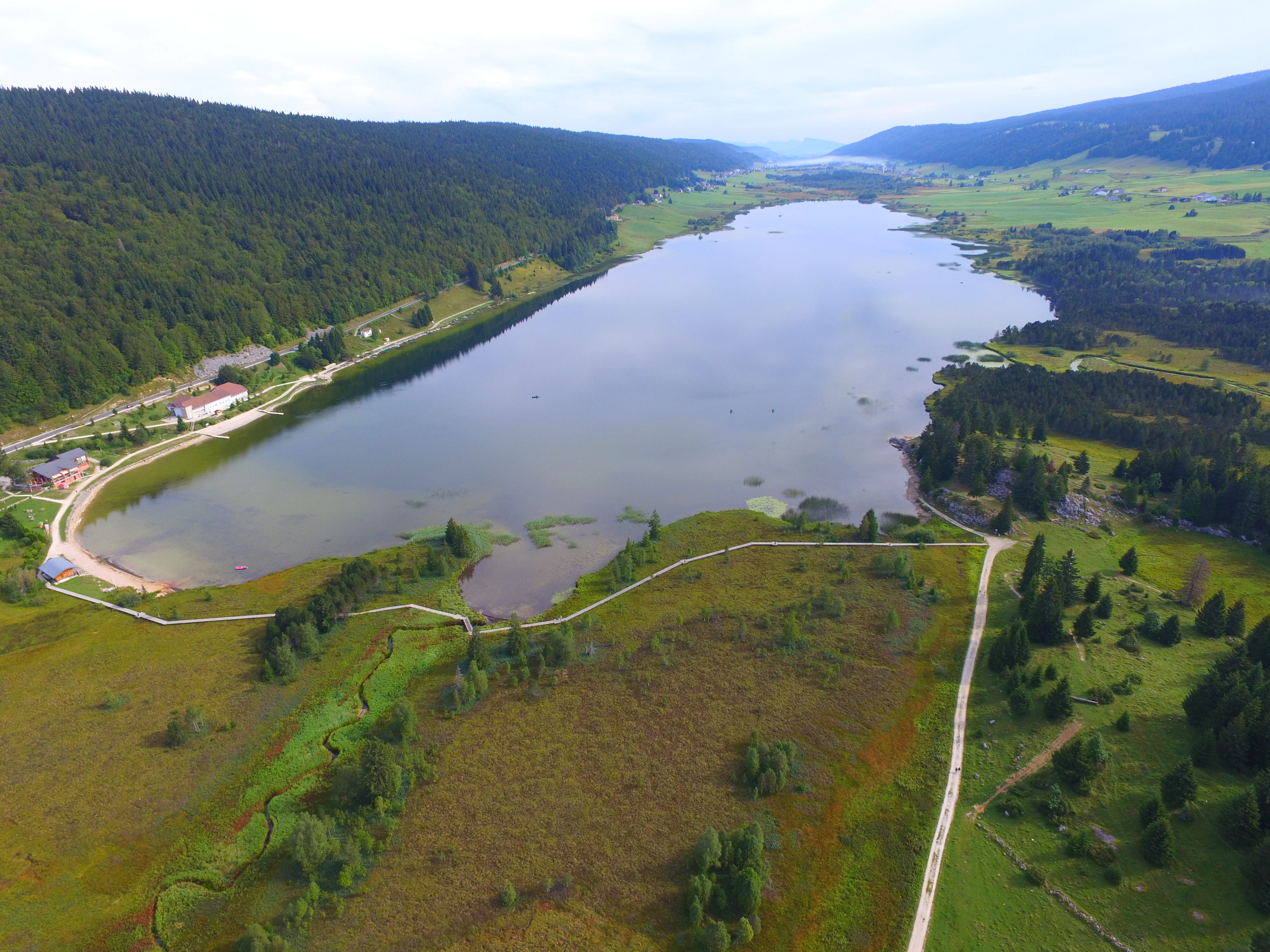 Una vista aérea de un lago de montaña alargado rodeado de densos bosques verdes y humedales en el macizo del Jura.