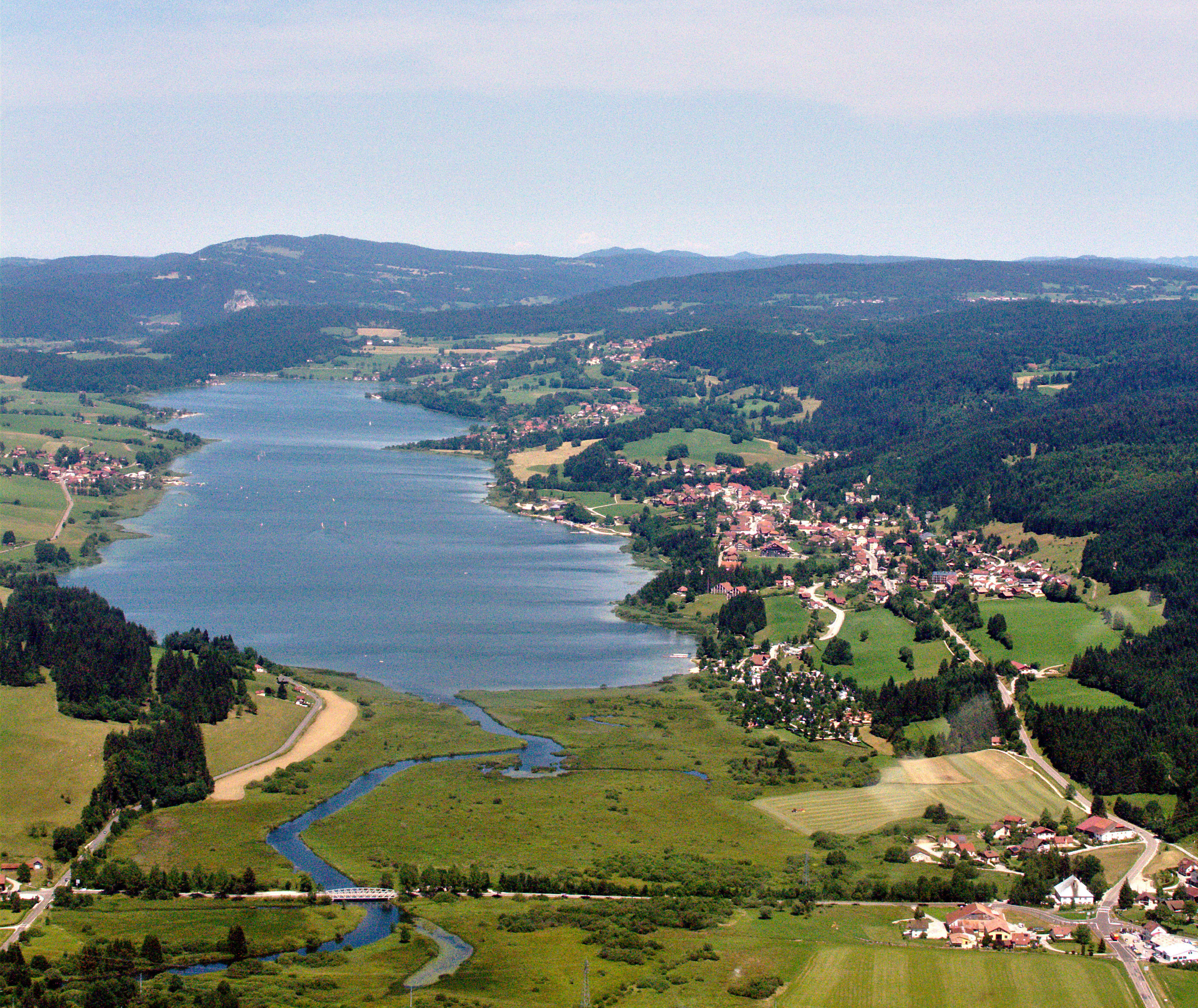 Una vista aérea que muestra un largo lago azul que se extiende a través de un valle en la cordillera del Jura, rodeado de campos verdes, bosques densos y pequeños asentamientos de aldeas.