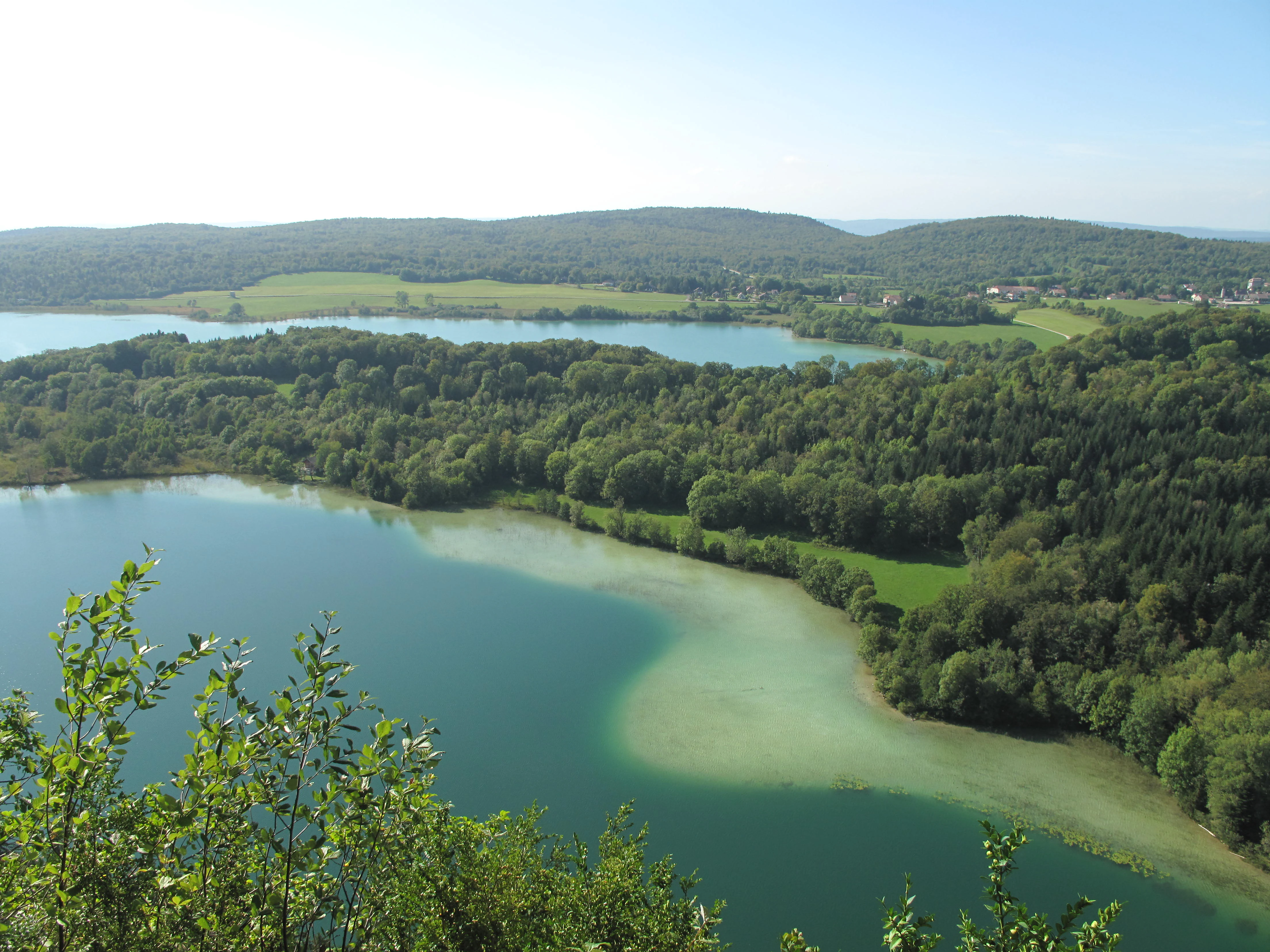 Vista en ángulo alto de dos lagos naturales serenos rodeados de denso bosque verde y colinas onduladas bajo un cielo brillante.