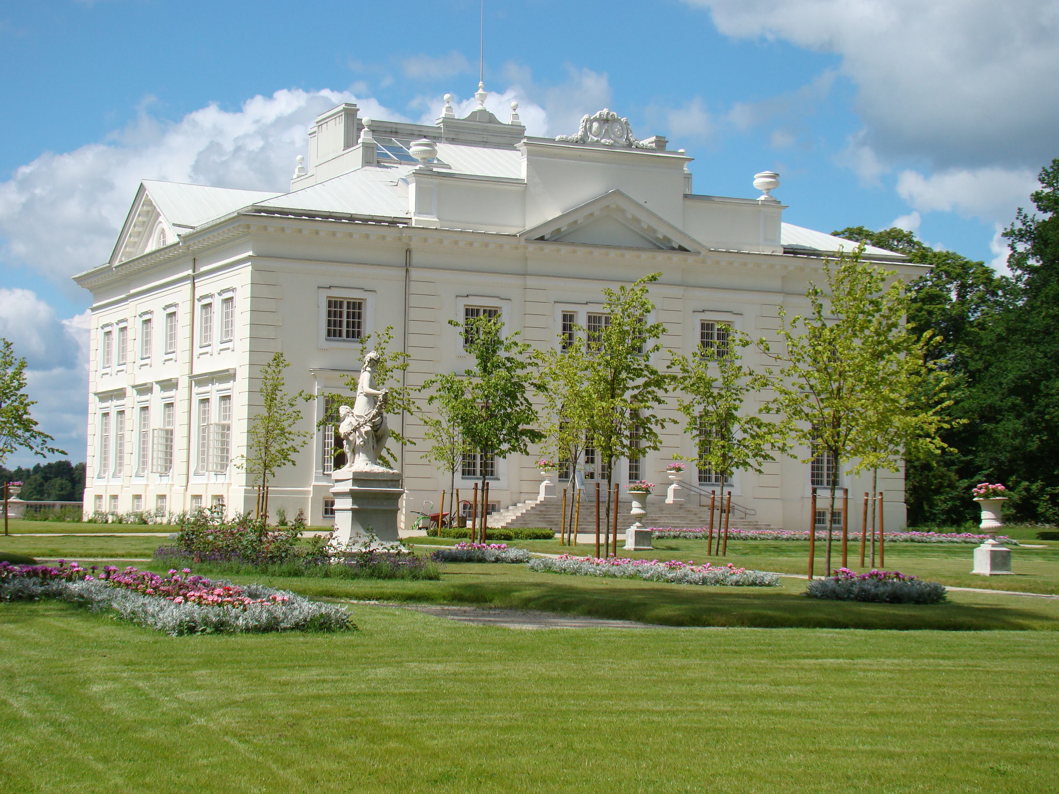 A large white manor house with a central statue in a landscaped garden, under a partly cloudy sky