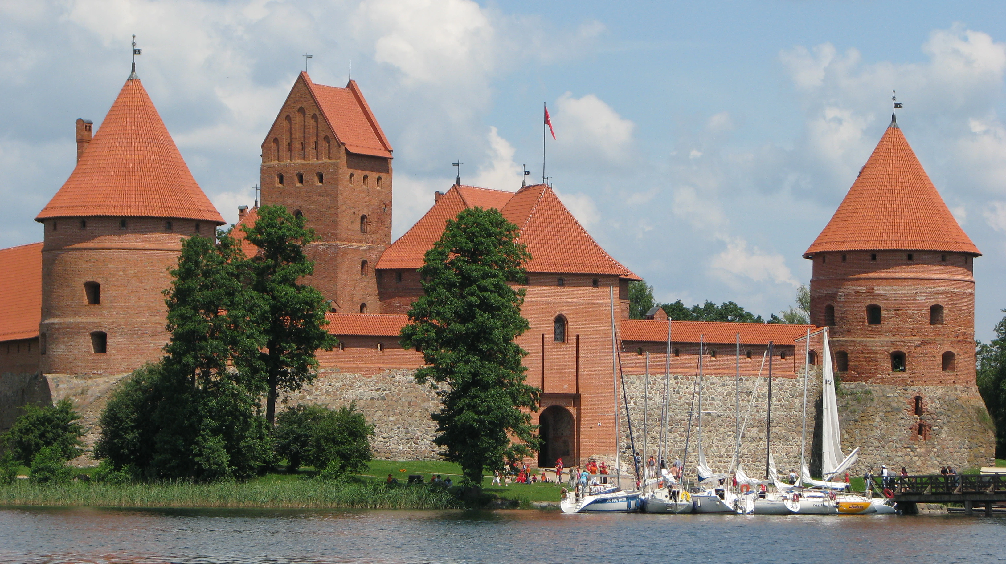 Red-roofed stone castle with multiple towers on a lake, surrounded by trees, with sailboats docked nearby