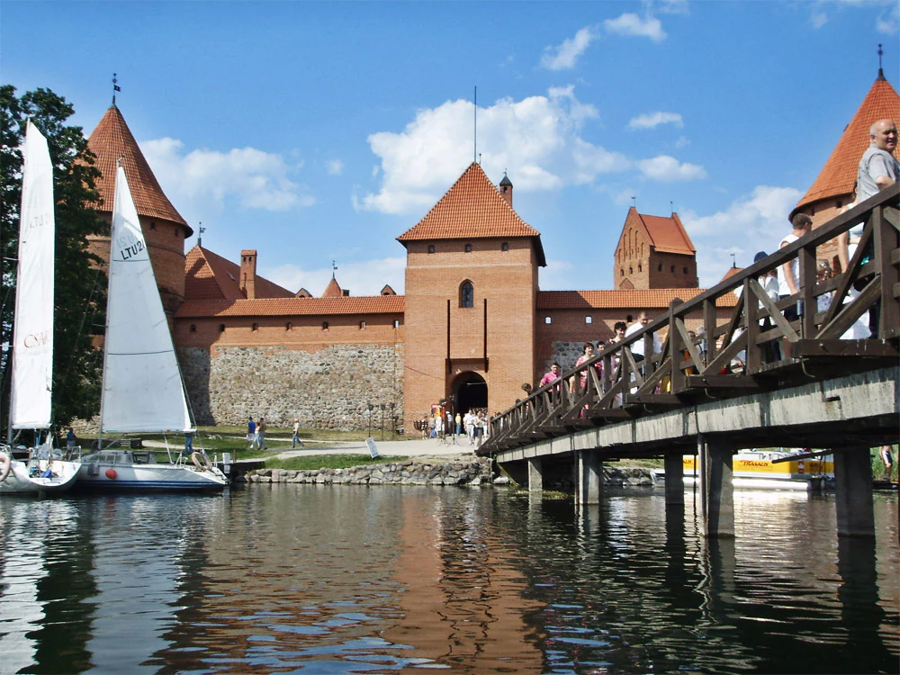 Trakai Castle with red-tiled roofs and stone walls, viewed across a body of water with a wooden bridge and sailboats