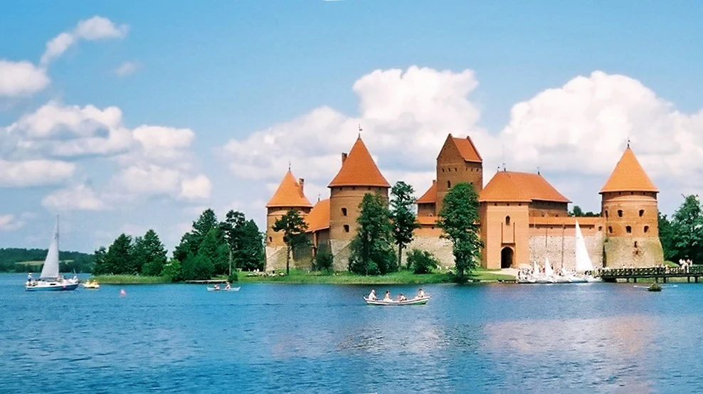 Trakai Island Castle with red roofs on a lake, surrounded by trees, with sailboats and a small boat on the water under a partly cloudy sky
