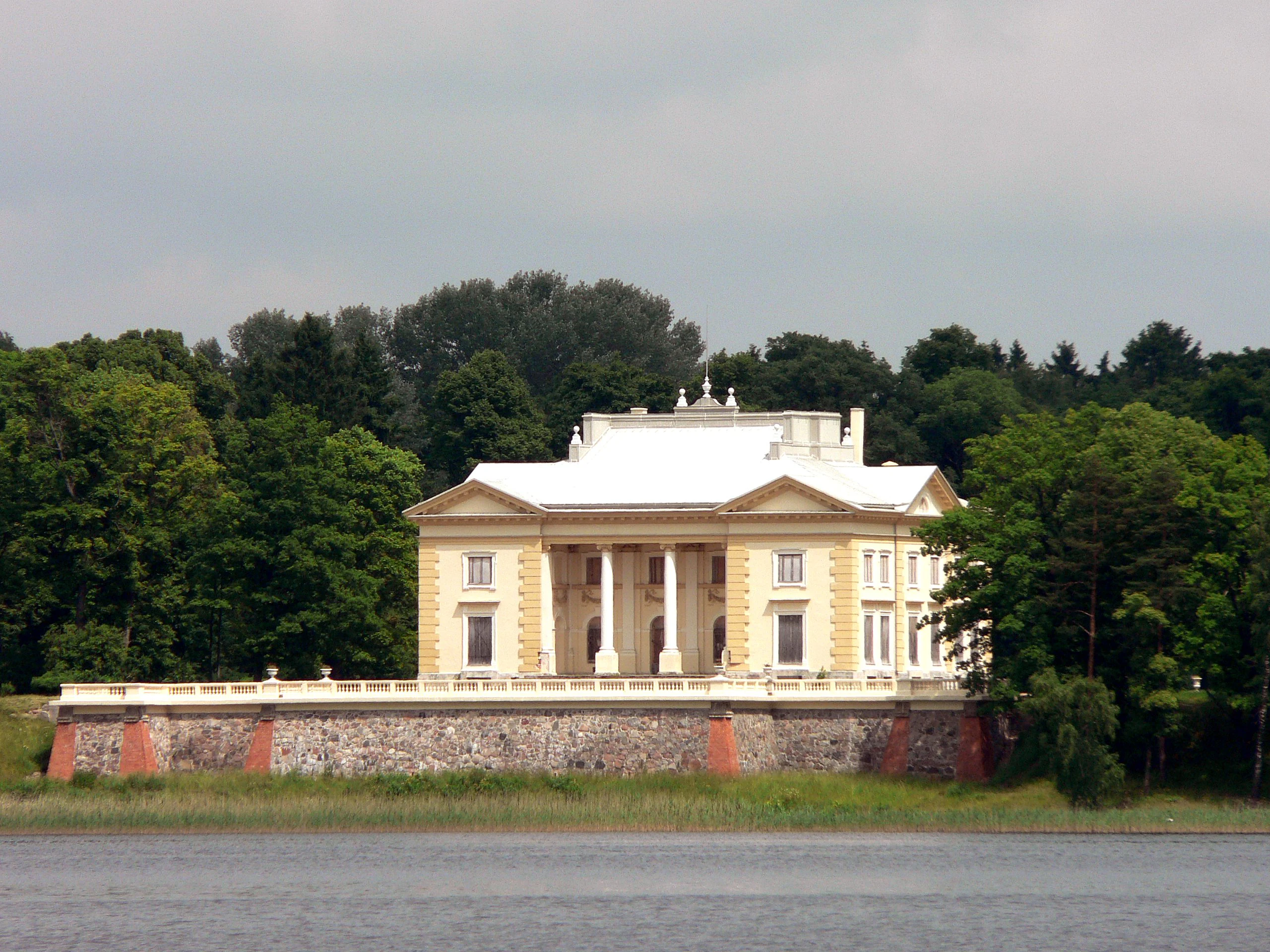 A large classical-style palace with columns on a stone foundation, situated by a body of water with green trees in the background