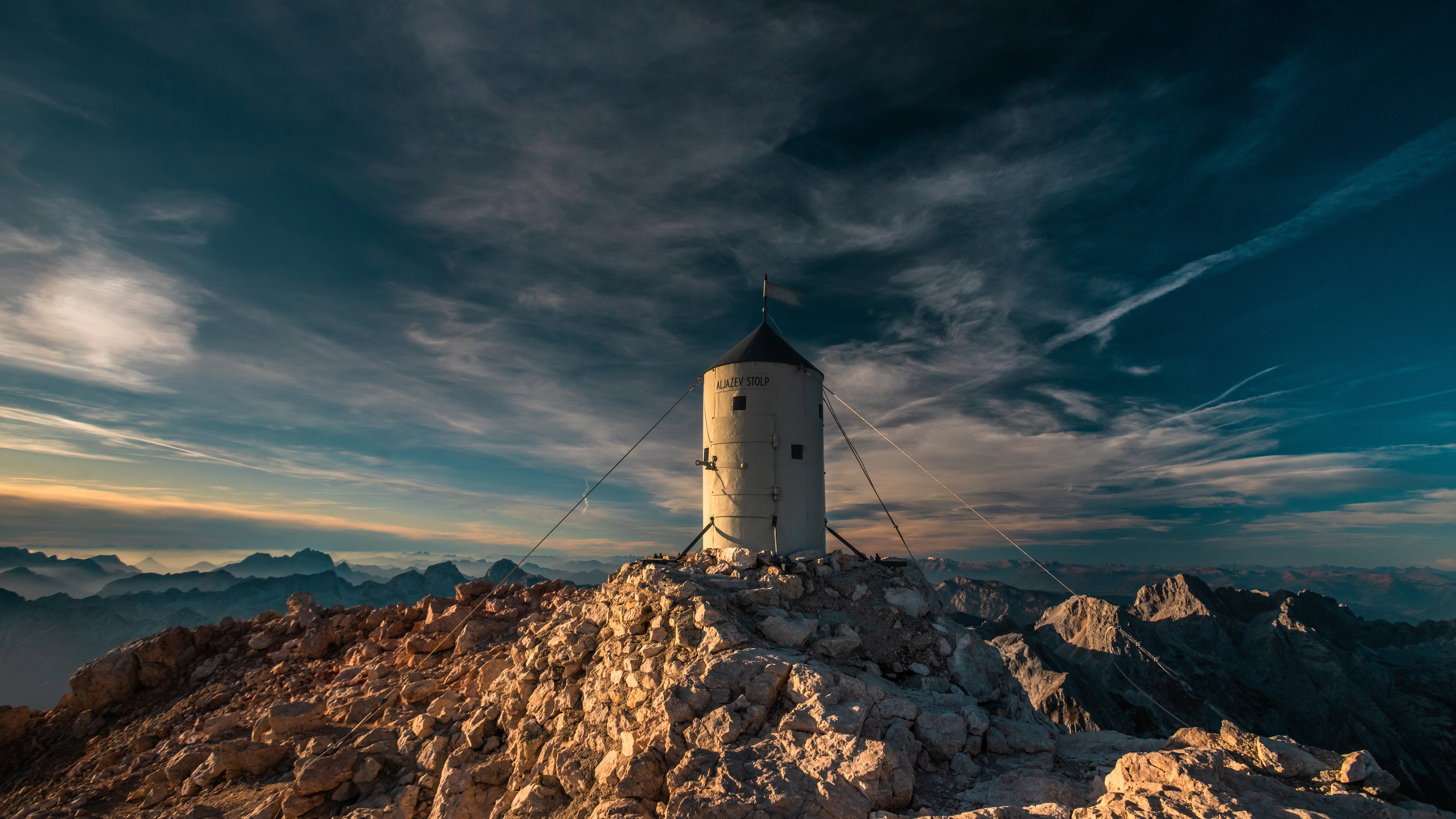 Aljaž Tower on a rocky mountain summit under a dramatic sky with distant peaks