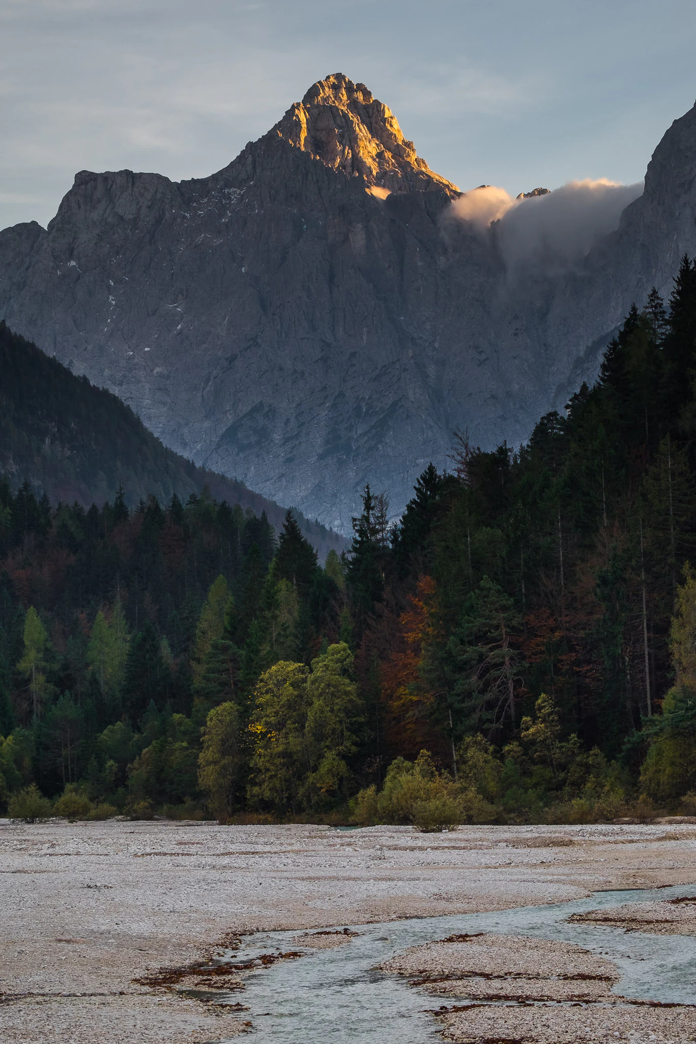 A river flows through a gravelly bed surrounded by dense forest with a mountain peak in the background under a cloudy sky