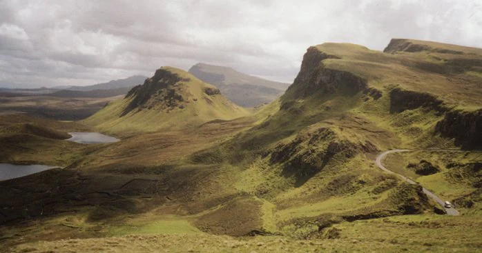 A wide view of the Quiraing landscape on the Isle of Skye, featuring craggy green hills, steep slopes, small bodies of water, and a narrow winding road.