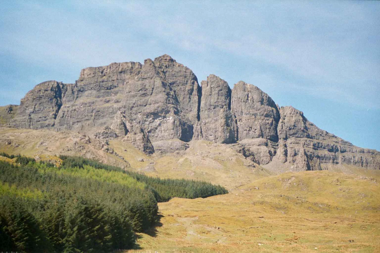 A wide, eye level landscape shot of a craggy, grey basalt rock face rising above a grassy hillside and a dense coniferous forest, under a pale blue sky.