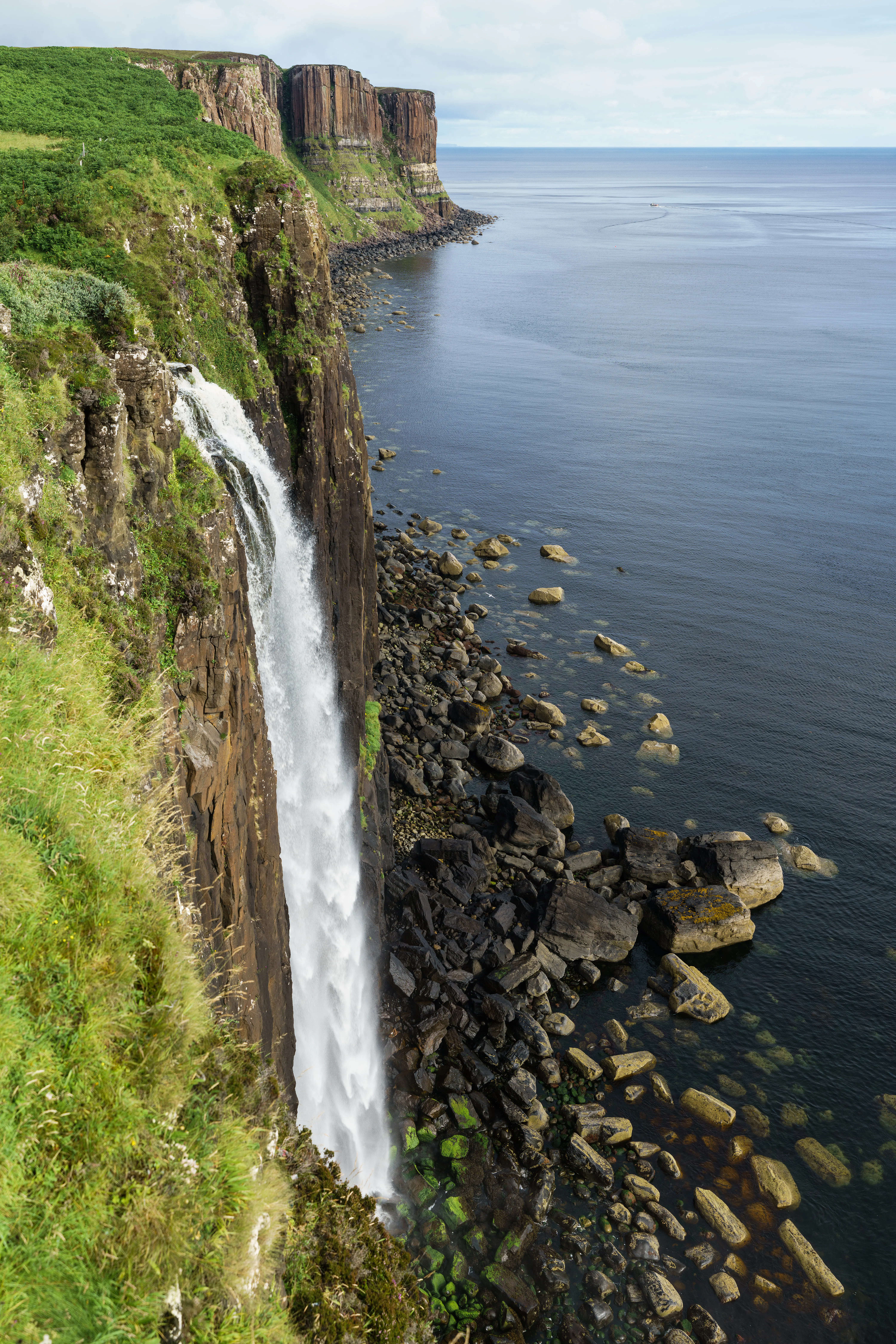 A high angle view of a narrow waterfall plunging from the edge of tall, dark basalt cliffs into the sea below.