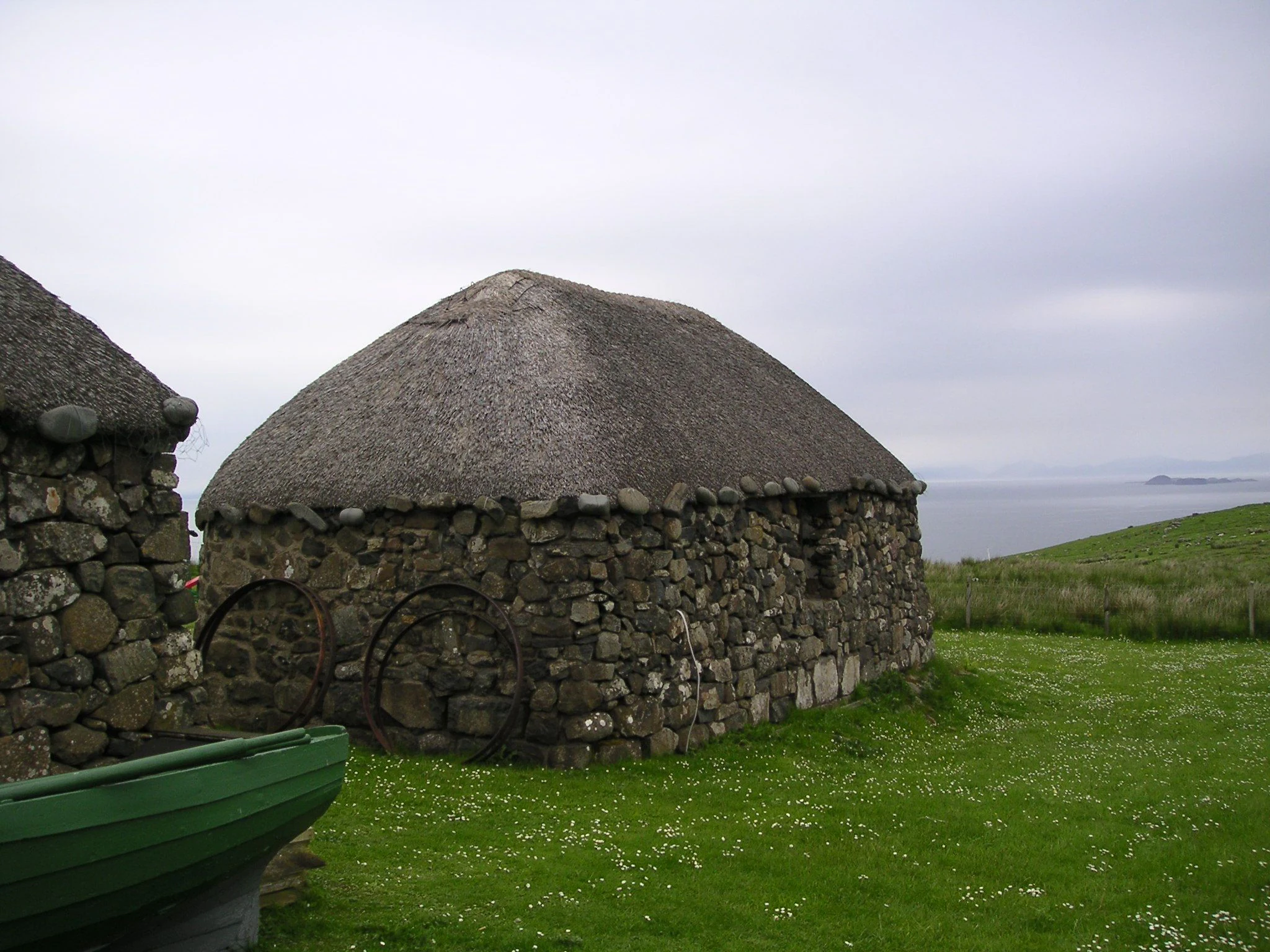 A traditional stone building with a thick thatched roof set in a grassy field overlooking the sea, with a green boat partially visible in the foreground.