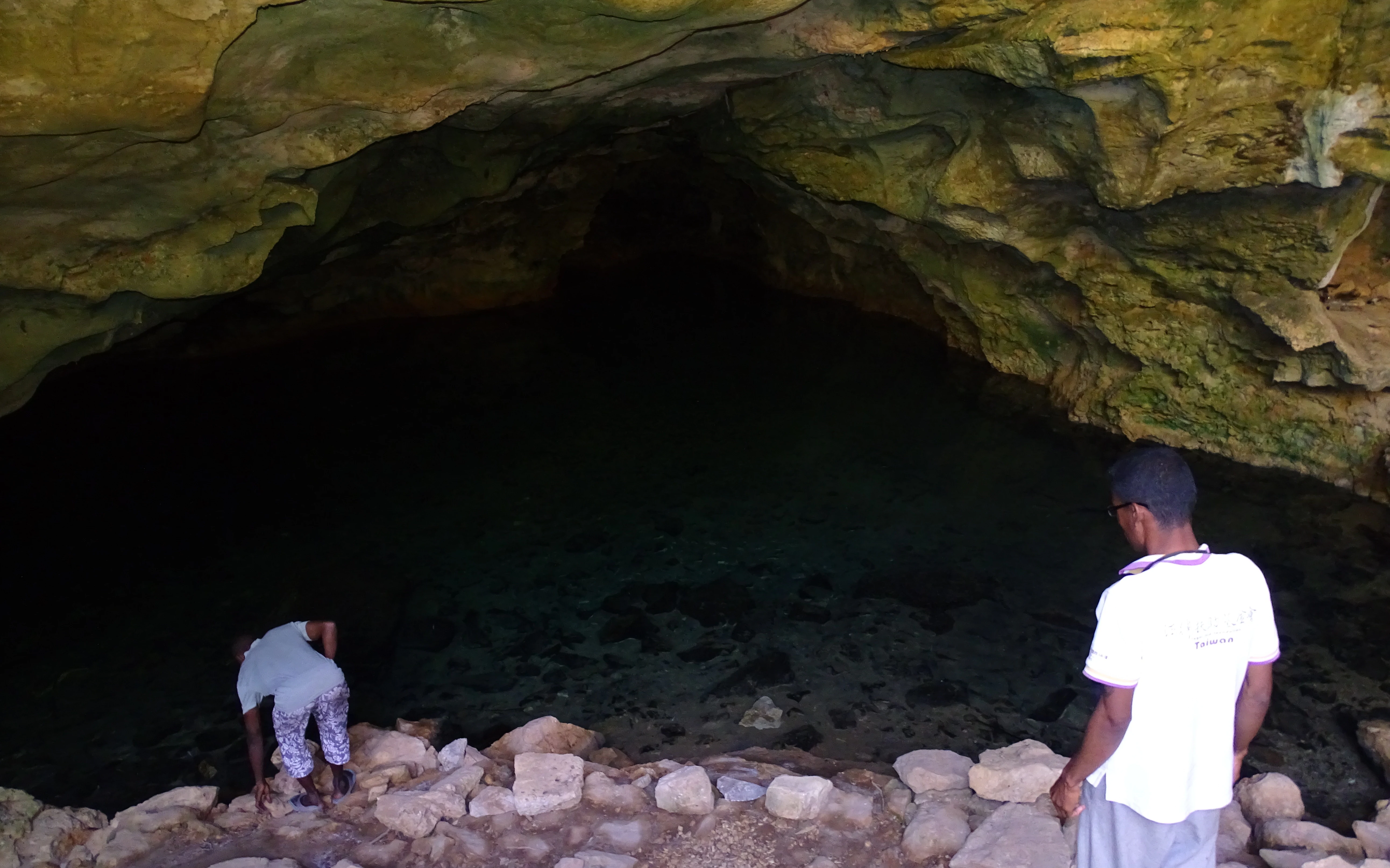 Two people standing at the edge of a water-filled limestone cave opening, looking into the dark cavern.
