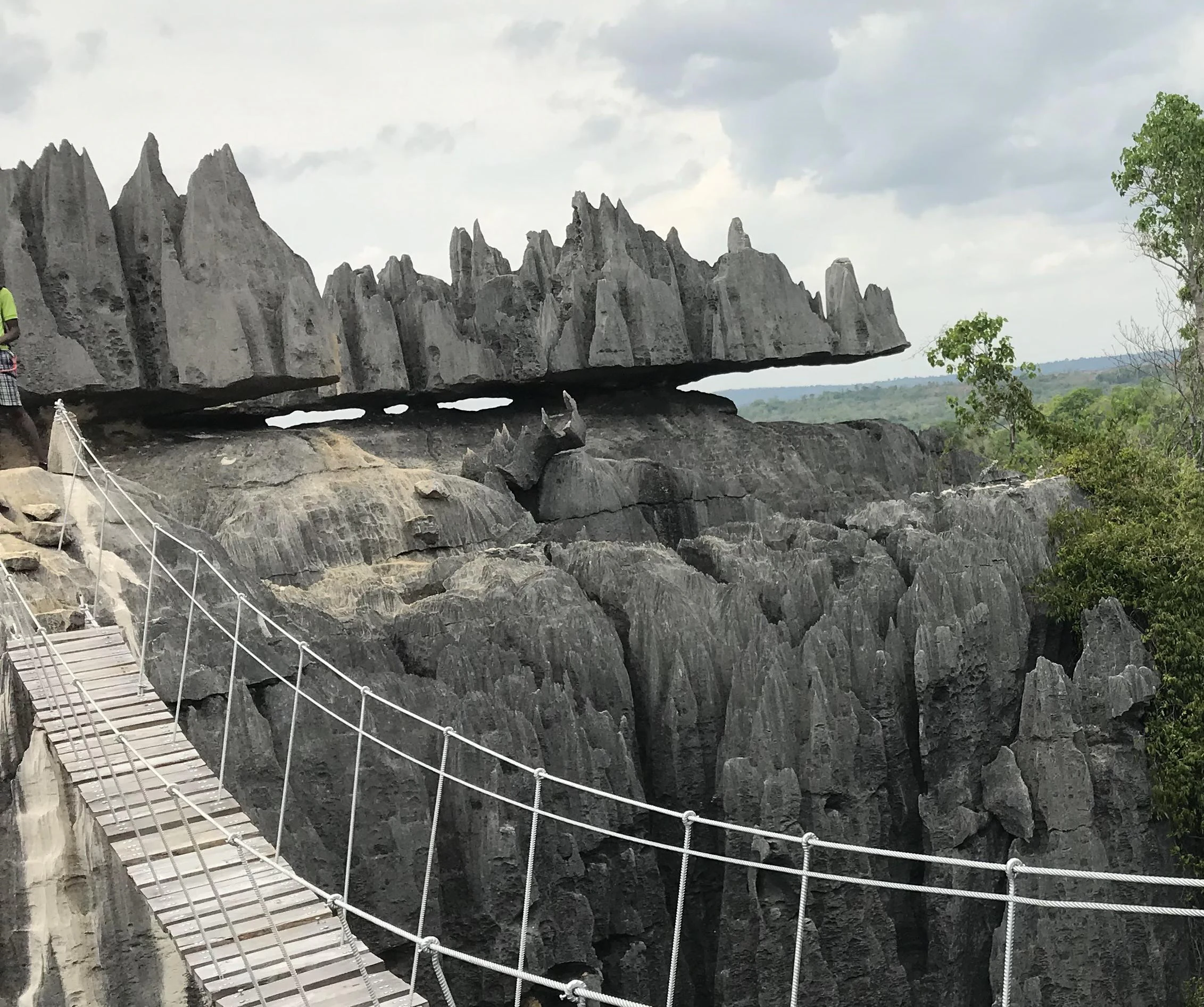 A wooden suspension bridge with metal cable railings leads towards jagged, grey limestone rock formations under a cloudy sky.
