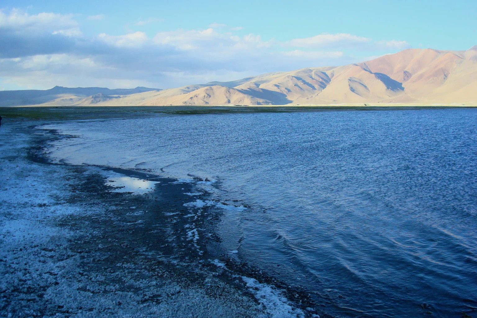 A wide view of a large, blue high-altitude salt lake with a salt-white shoreline and dry, rugged mountains in the distance under a clear sky.